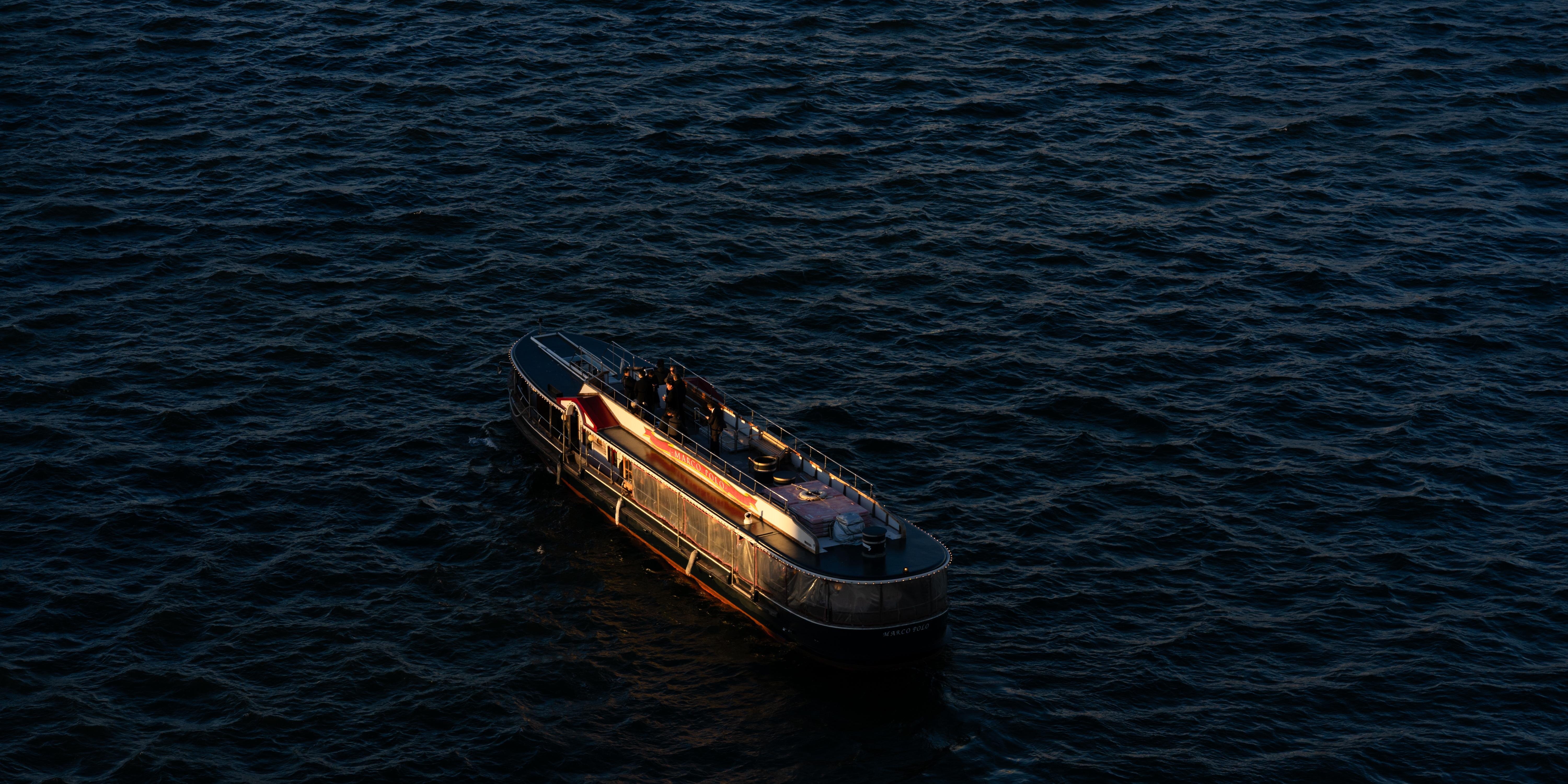 A small boat with a dark hull and an illuminated interior floats on a dark, textured water surface. The boat has several people on board and features a rounded front and an open area on top. Soft warm lighting highlights the boat’s details, contrasting with the surrounding deep blue water.