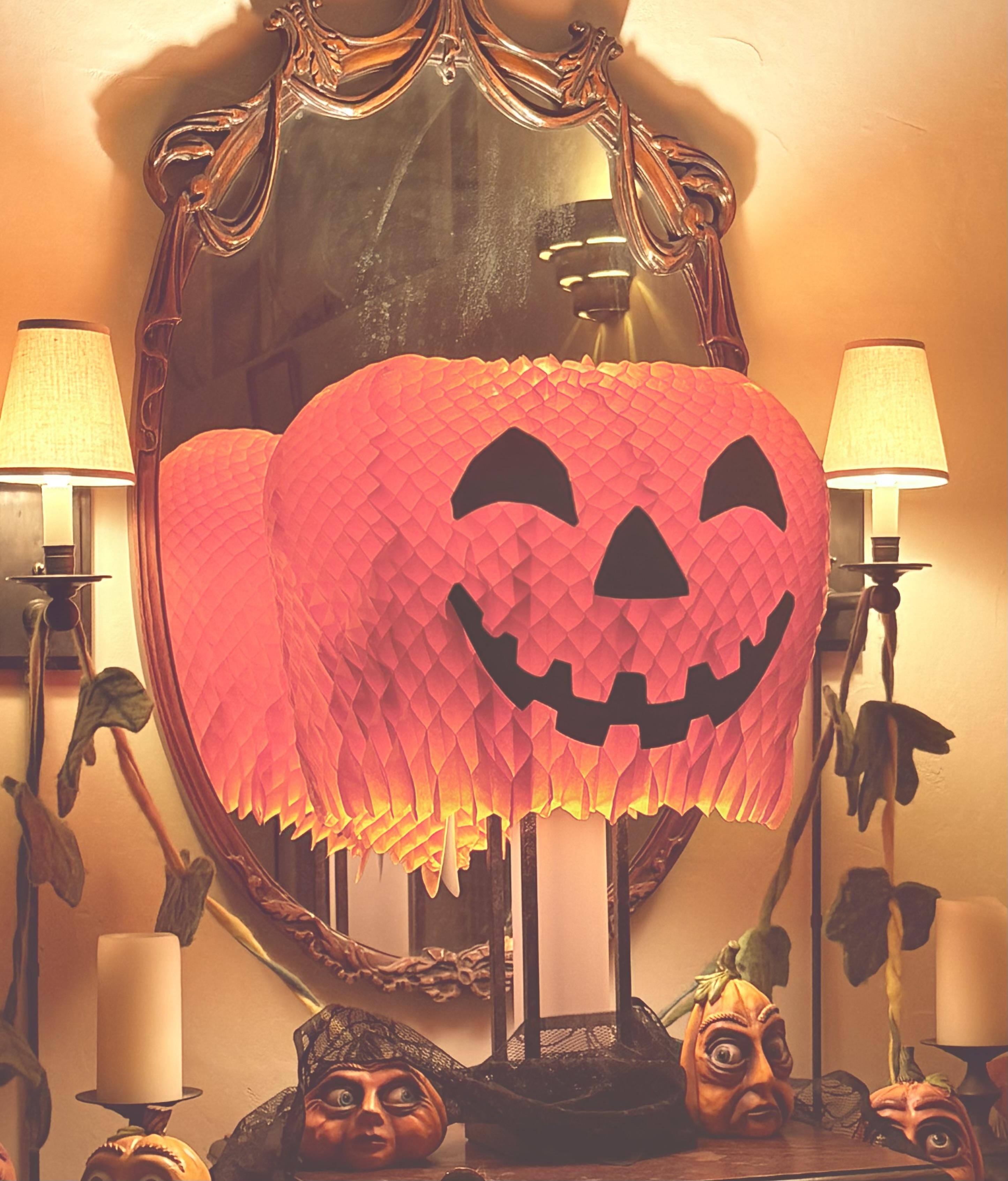 A large, orange, paper pumpkin lantern with a black smiling face sits in front of an ornate mirror, creating a reflective effect. Two table lamps with yellow shades flank the mirror. Below the lantern, on a wooden surface, are decorative pumpkin masks with carved faces and green leaves, and two unlit candles in holders.