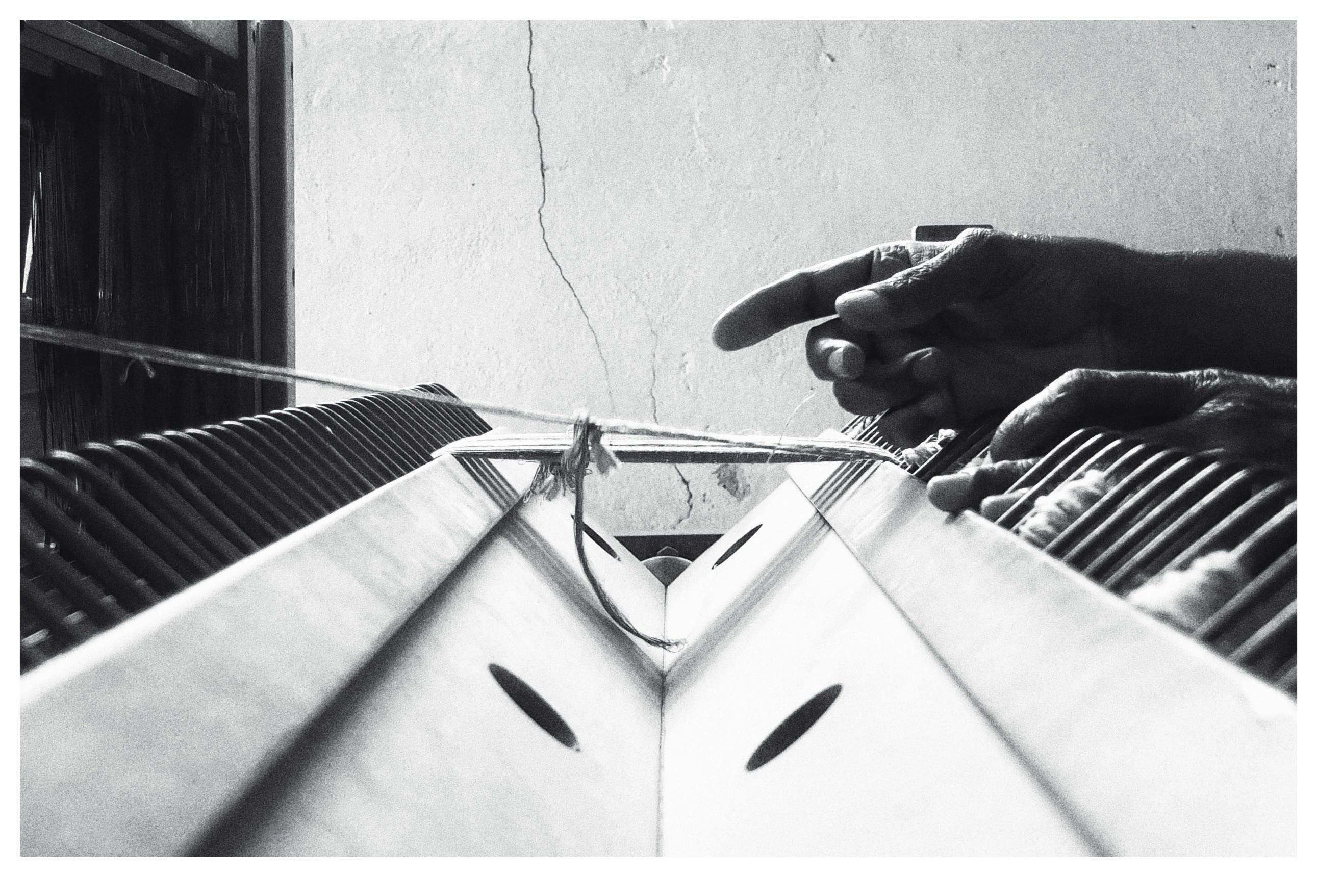 Black-and-white close-up of hands operating a traditional loom, with taut threads stretched across the frame and rows of wooden slats and heddles on both sides; a cracked plaster wall in the background.