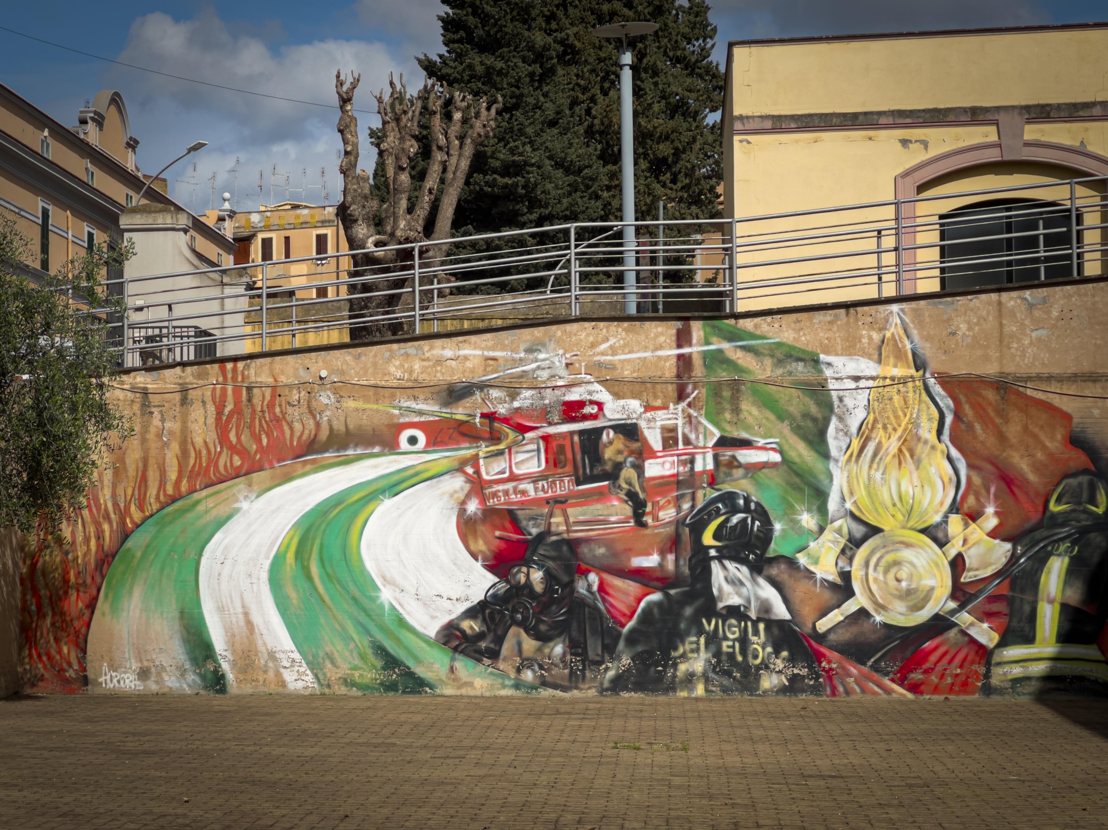 Murale che raffigura una scena di vigili del fuoco in azione. L'opera d'arte include pompieri in azione, con uno che indossa un equipaggiamento etichettato "Vigili del Fuoco." È presente un elicottero antincendio che vola sopra, e lo sfondo include fiamme e una rappresentazione della bandiera italiana. Il murale è realizzato su un muro esterno, con colori vivaci e immagini dinamiche.