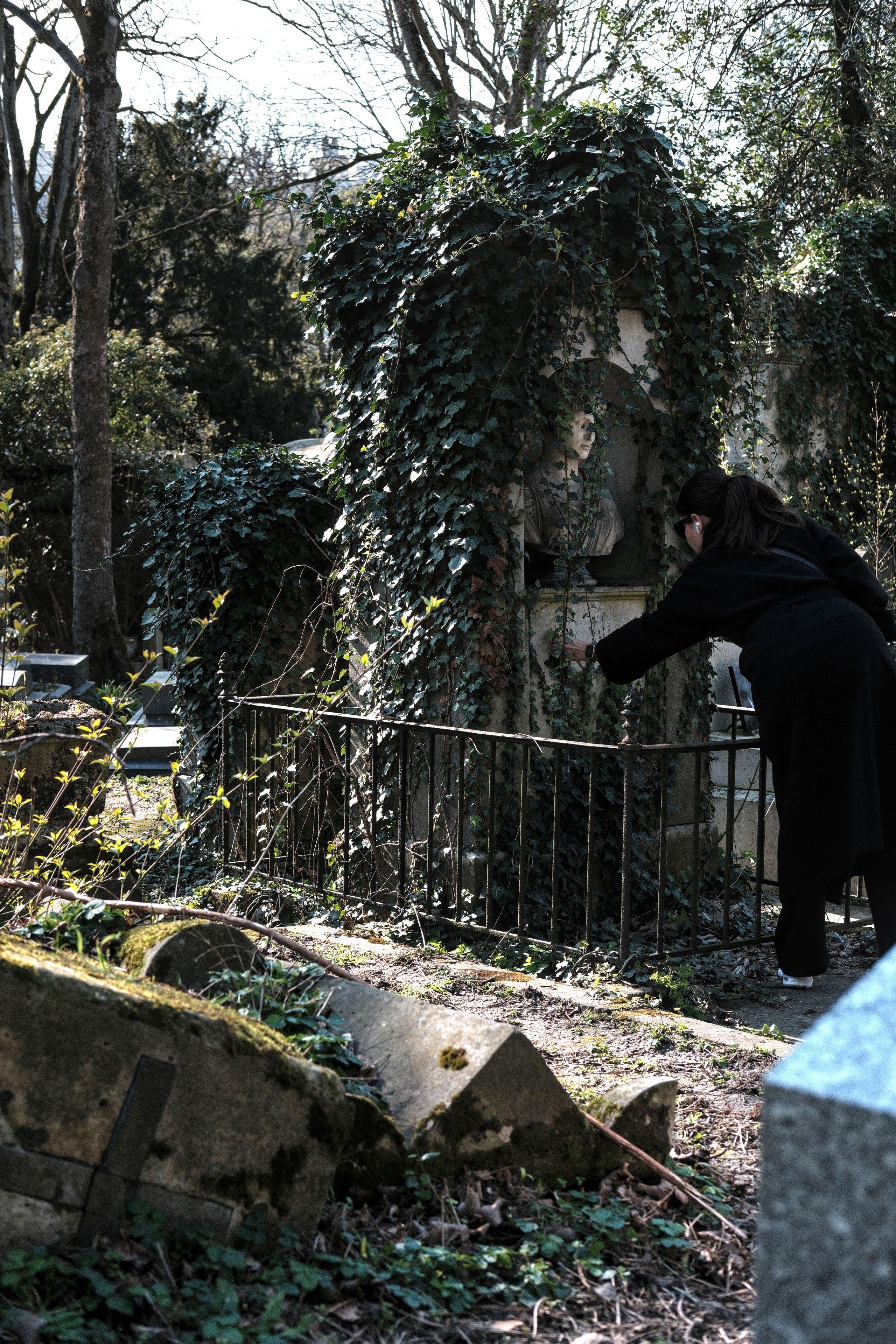 Photo verticale. Une vieille tombe protégée par une grille et couverte de lierre sur le dessus. Un buste se trouve sous le lierre et une femme en manteau noir est en train d’enlever du lierre de la plaque sous le buste