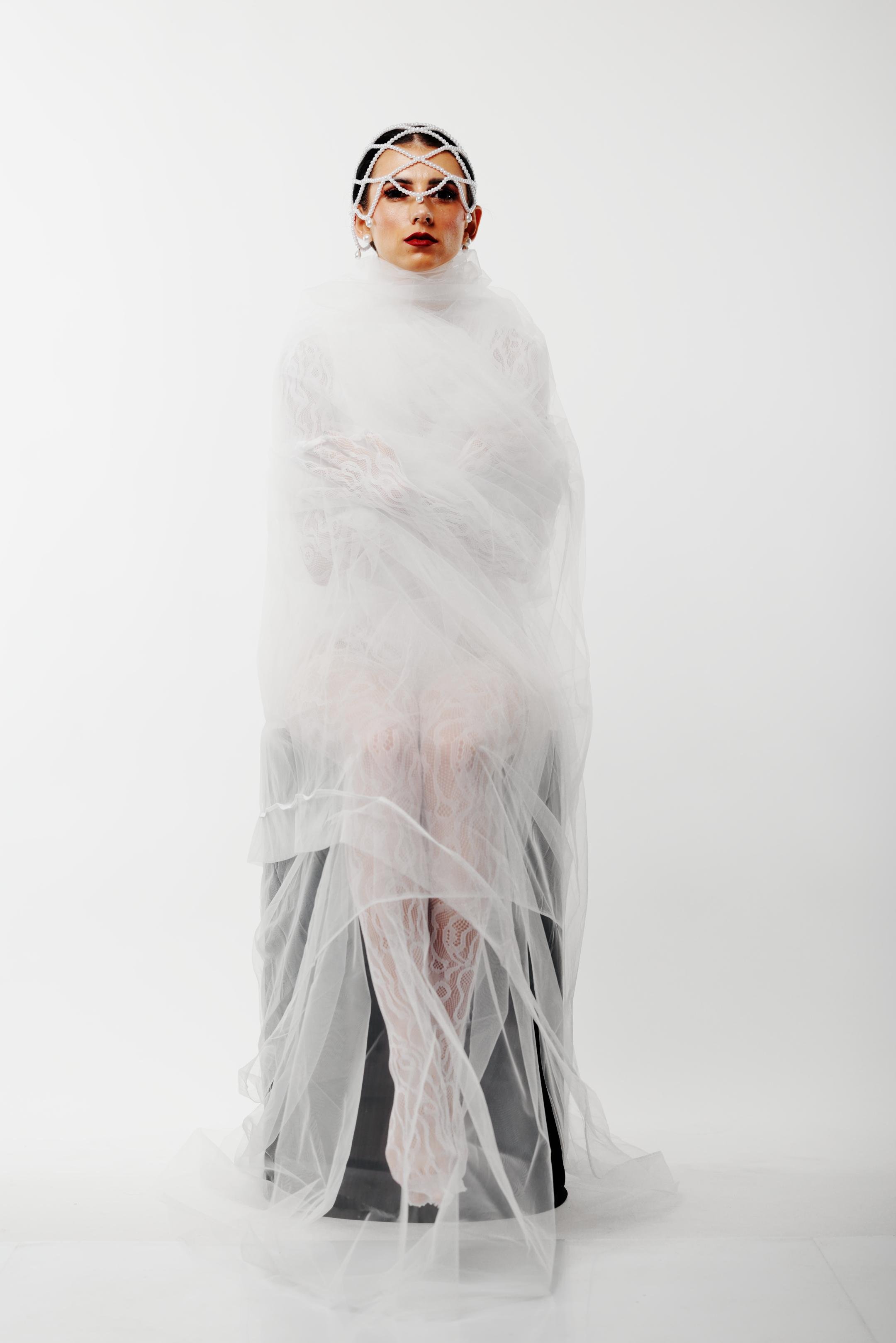 Person seated against a white background, wrapped in layers of translucent white fabric. Wearing a pearl-like headpiece over the forehead and makeup with dark lipstick. White lace is slightly visible through the fabric.