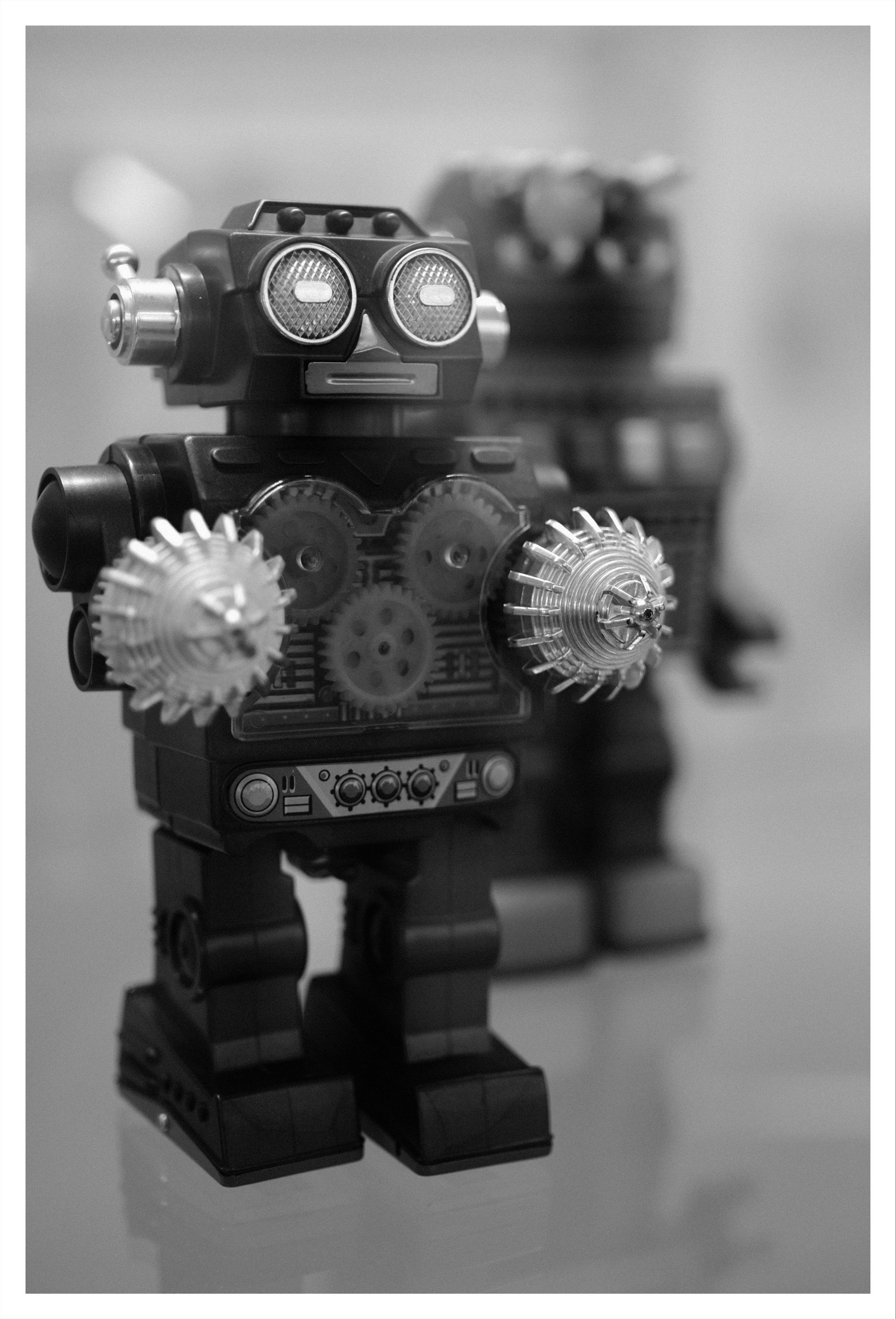 Black-and-white close-up of a vintage toy robot with large circular eyes and two spiked disc-shaped hands; a second robot stands blurred in the background on a smooth reflective surface.