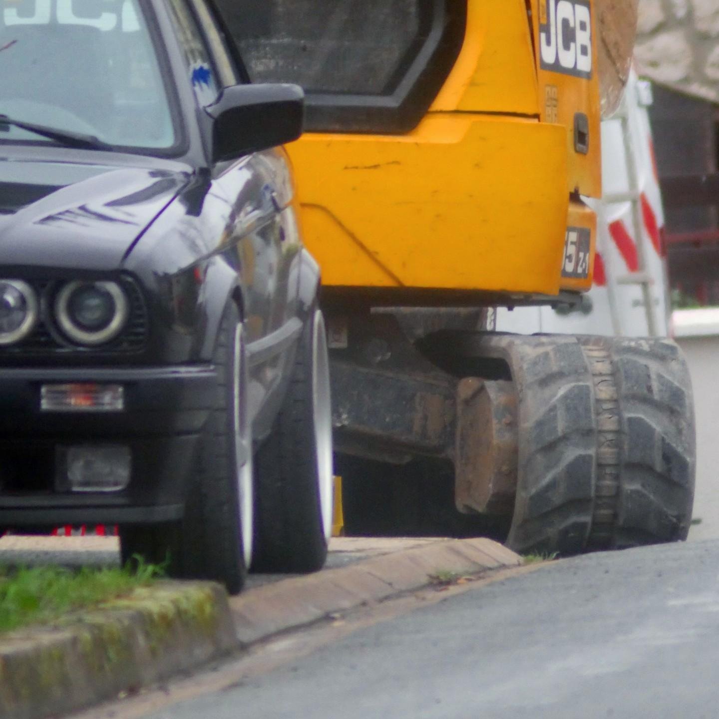 The image shows a black car parked closely in front of a large yellow construction vehicle, which appears to be a JCB machine. They are positioned near a roadside curb.