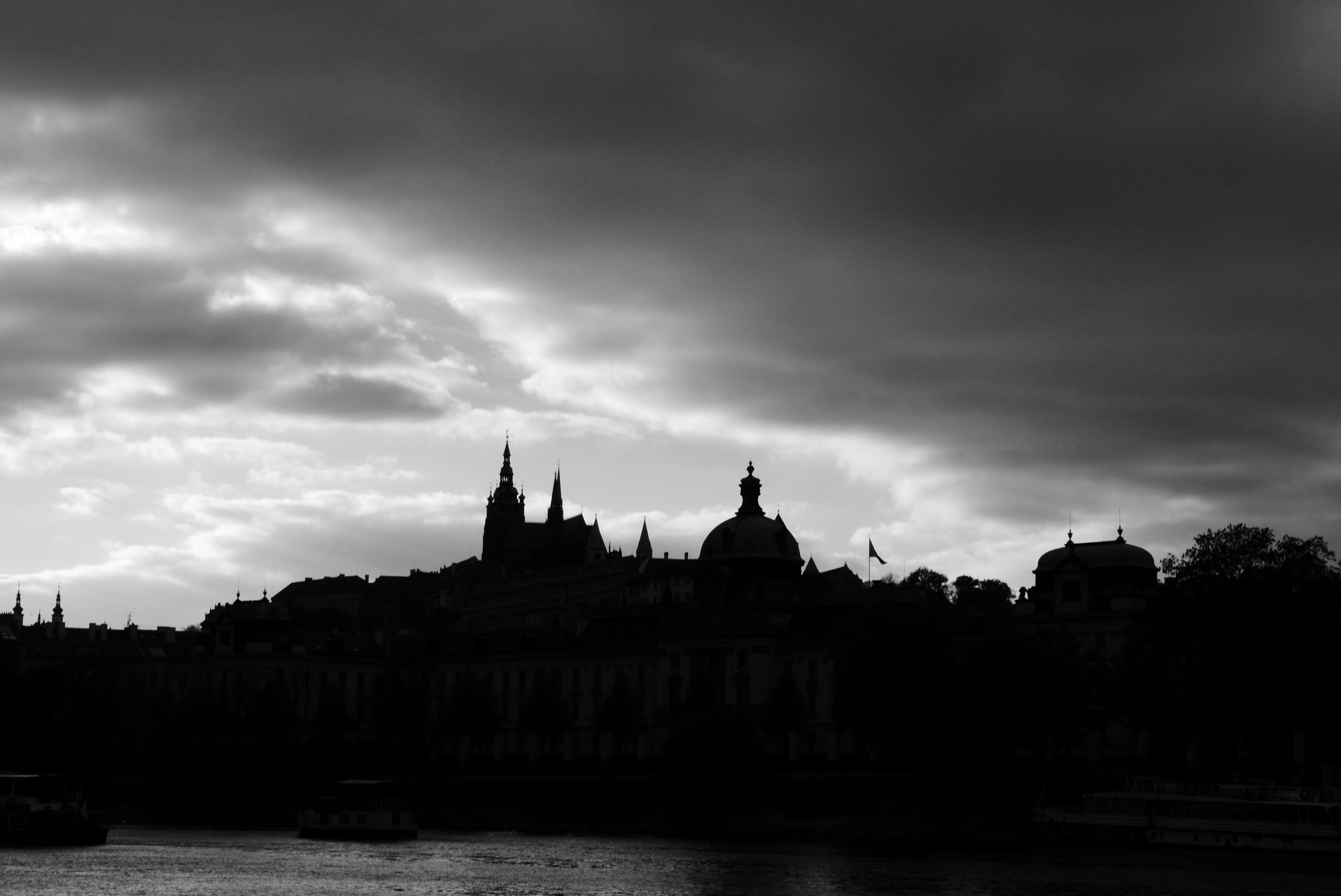 This image shows a dramatic black and white silhouette of a cityscape, likely featuring historical architecture. The skyline includes spires and domed structures, possibly a cathedral or castle. The scene is contrasted against a cloudy sky, adding to the dramatic effect. There also appears to be a river or body of water in the foreground.