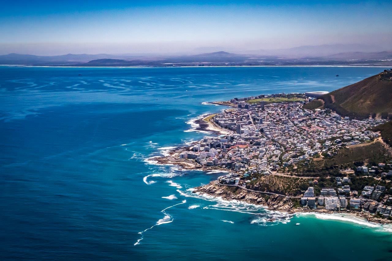 This image shows an aerial view of a coastal city with prominent blue ocean waters and a clear skyline. There's a mixture of urban development with buildings clustered near the coastline. The land juts into the ocean, forming a distinctive peninsula. It's a clear day with mountain ranges visible in the background.