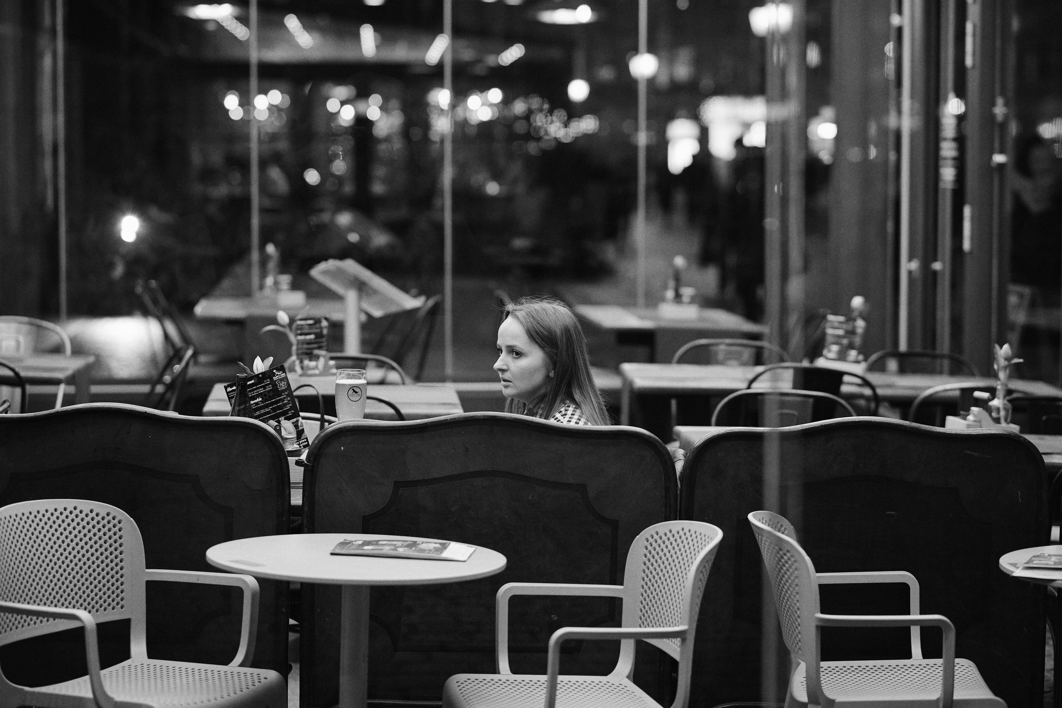 The image is in black and white, showing a cafe with tables and chairs. There is a woman seated indoors, looking to her right. The cafe has a modern, well-lit interior with large glass windows, and there are other tables and decor visible in the background.