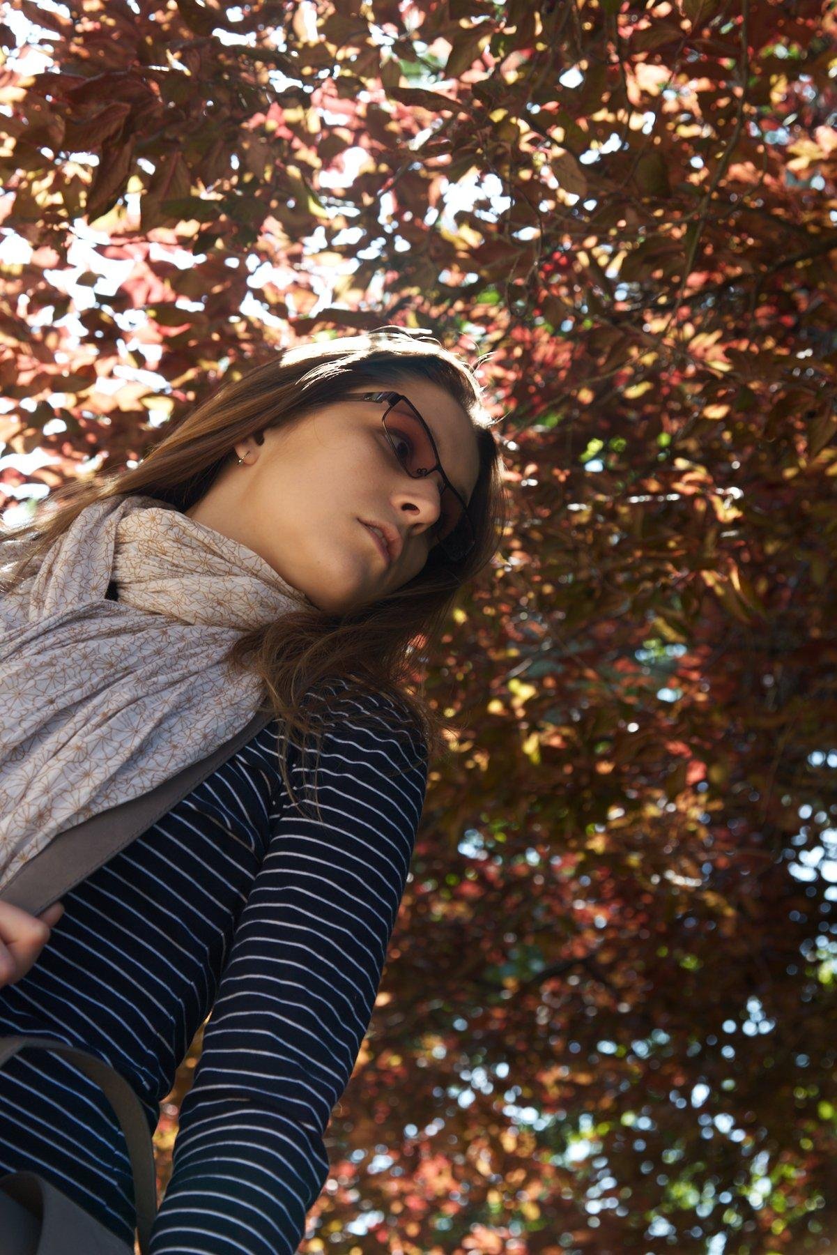 Photo format portrait en contre plongée montrant une jeune femme brune avec des lunettes auto tintantes, un haut bleu marine à rayures blanches horizontales, un foulard blanc au motifs floraux marron clair. Elle regarde vers la droite. Au dessus d’elle les feuilles marrons/orangées de l’arbre forme le fond de l’image