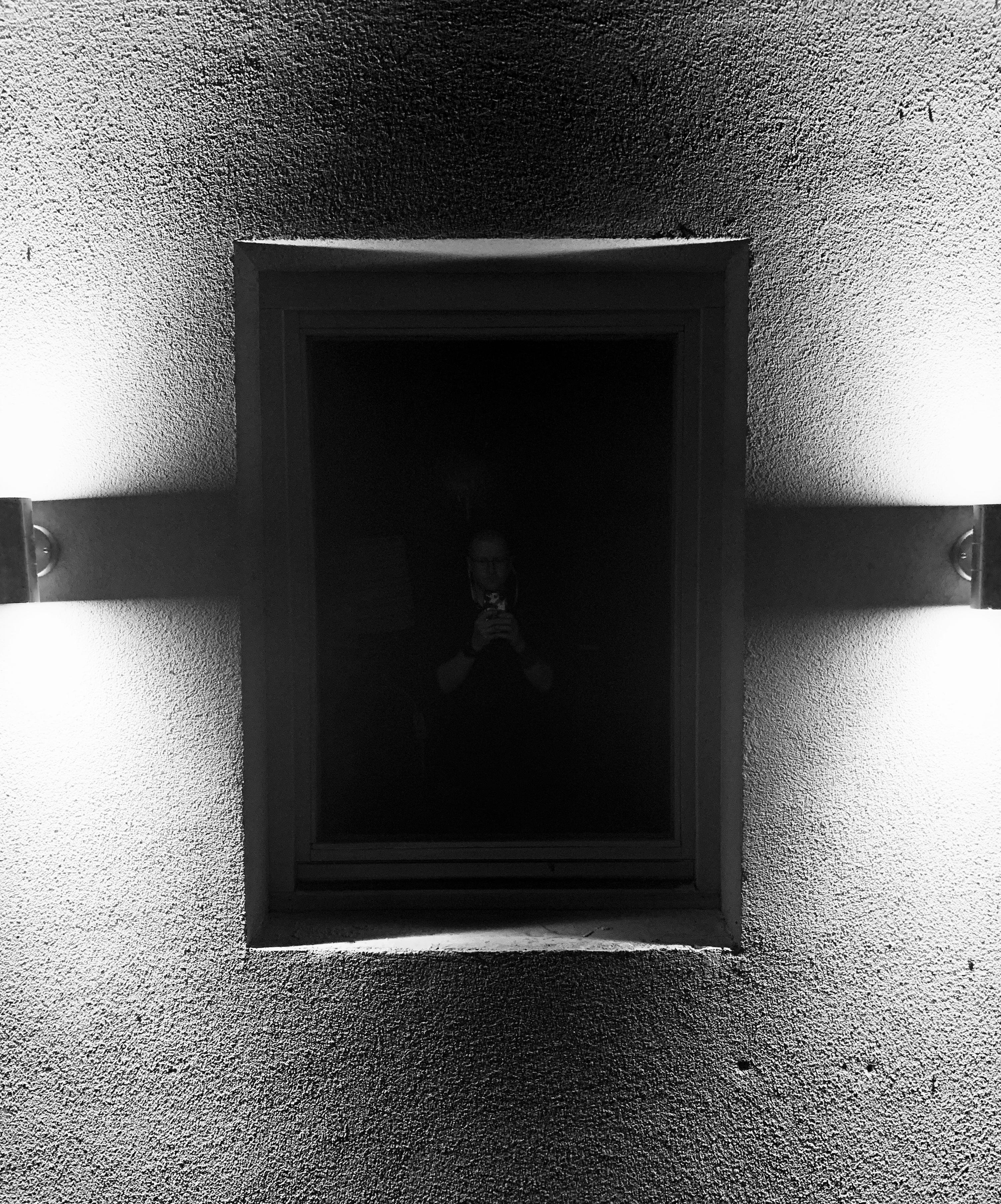 Black and white photo with a wall, lights, window in the center and a reflection of a photographer.