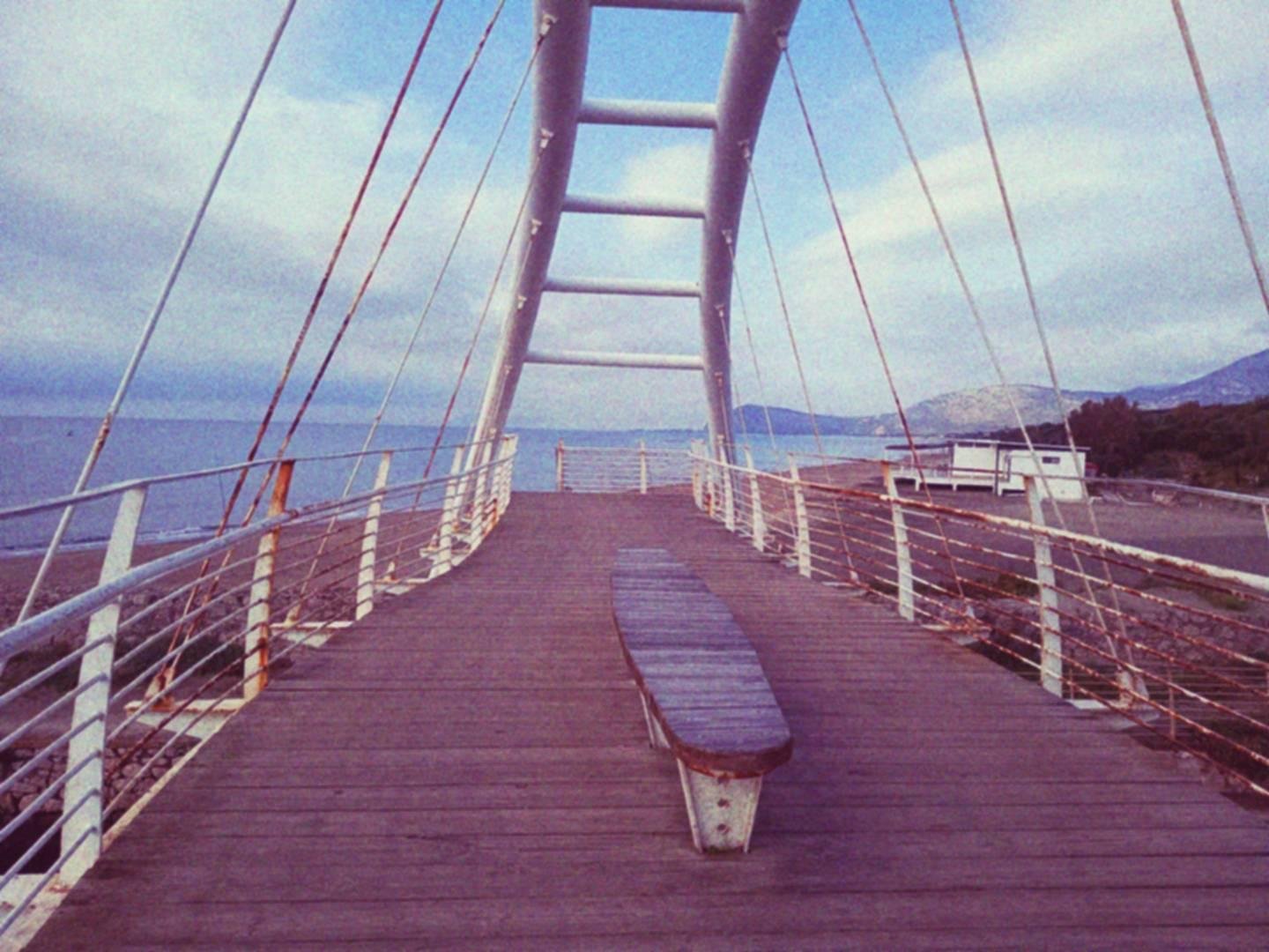 Ponte in legno e metallo che collega le due sponde di uno stretto canale. Un arco metallico, rinforzato da tiranti, sovrasta la passerella in legno sulla quale è posta una panchina. Il cielo è nuvoloso, in lontananza si vede un promontorio.