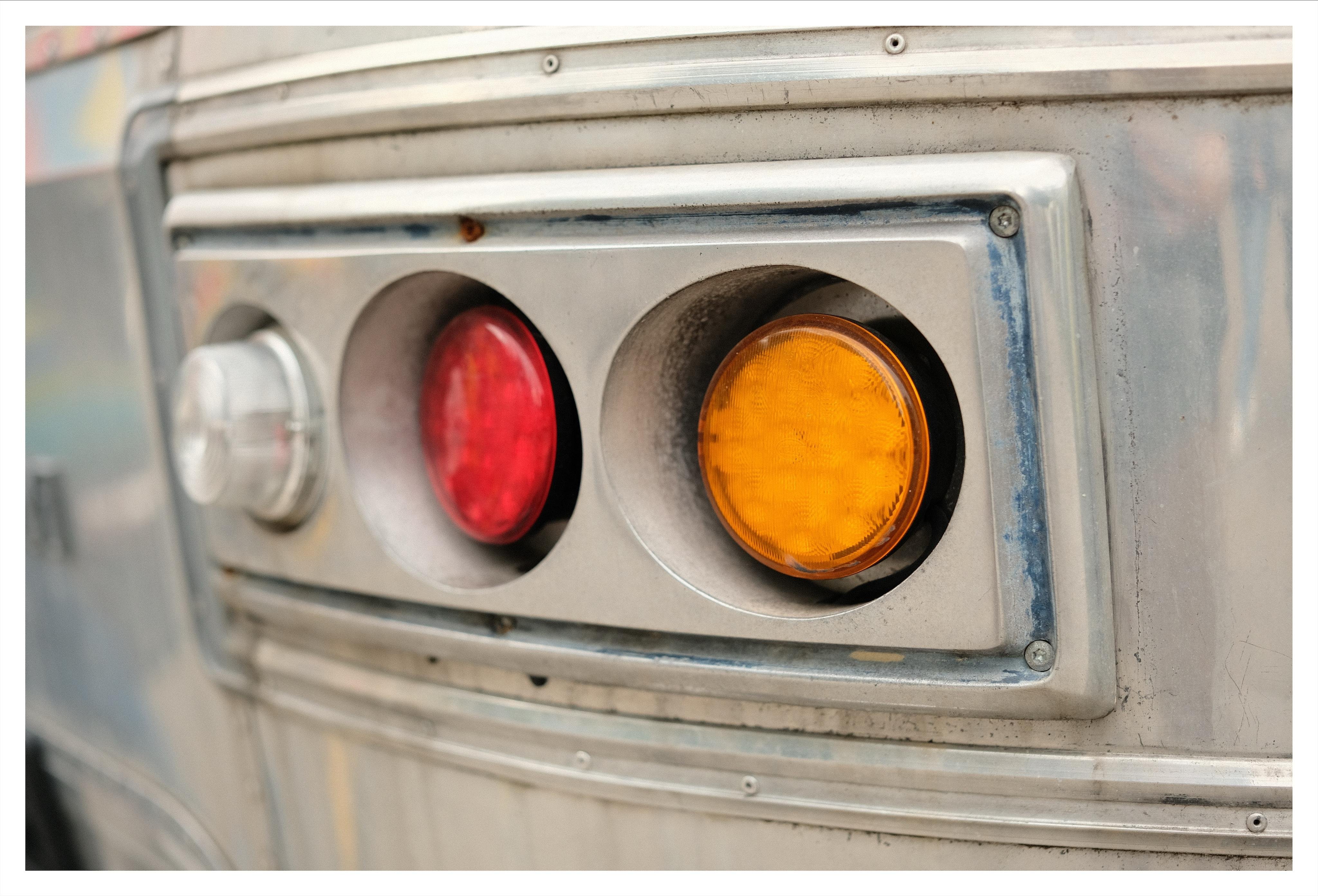 Close-up of a rectangular metal light panel on the silver trailer, featuring a red circular lens and an amber circular lens in recessed housings, with a small round metal cap on the left and visible screws and trim around the panel.
