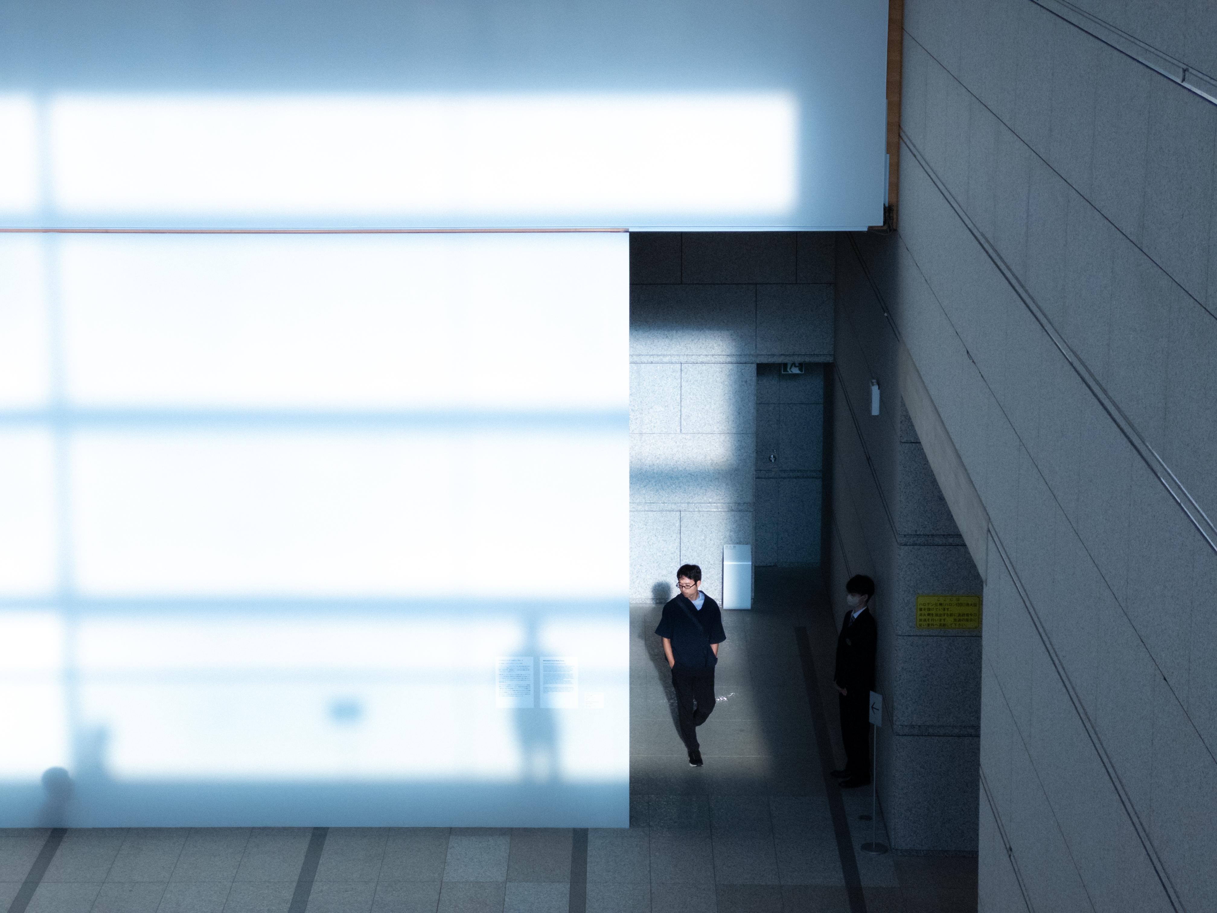 A person walks on a tiled floor through a narrow, shadowy corridor; sunlight filters through a large glass wall with a grid pattern, casting geometric shadows across the space. A second person, partially visible, stands in the shadow against a wall on the right. The setting features modern architecture with smooth stone surfaces.