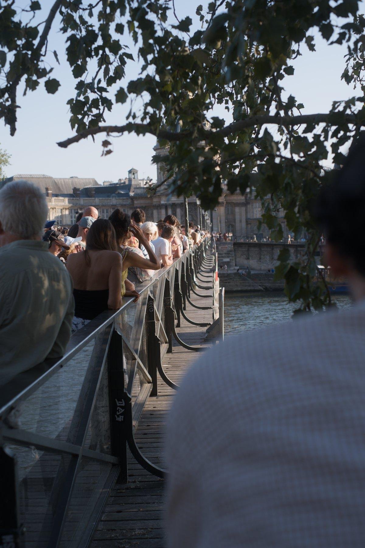 Photo verticale. Par dessus l’épaule d’une personne, on voit la barrière de sécurité du pont des arts côté Seine. Côté pont, des gens sont présent tout le long avec leur lunettes de protection pour regarder l’eclipse