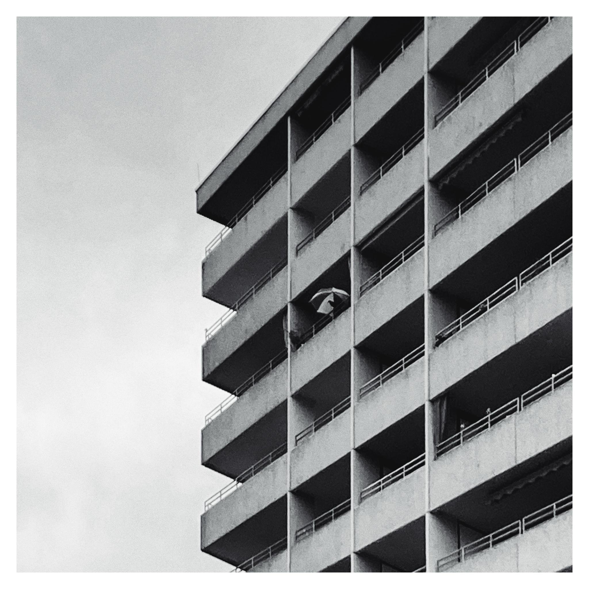 Black-and-white photo of a concrete apartment building with stacked balconies, viewed from below at an angle; a single open umbrella is visible on a mid-level balcony against an overcast sky.