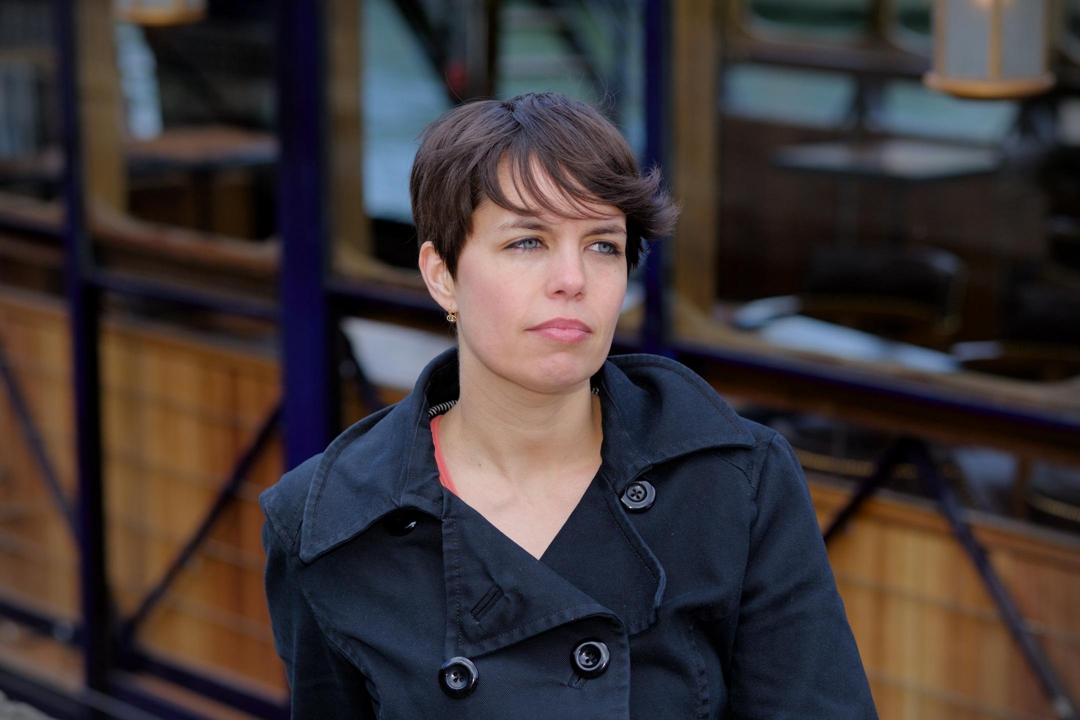 Une brune au centre de la photo, cheveux courts, le visage tourné de 3/4 regardant vers la droite du cadre avec des yeux perçant. Un bout de péniche avec du bois en fond