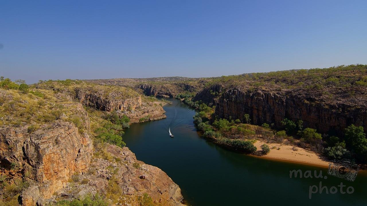 The image shows a scenic landscape featuring a river winding through a rocky canyon. The surrounding area is covered with sparse vegetation typical of a semi-arid environment. The sky is clear and blue, suggesting a sunny day. There is a boat visible on the river, and the canyon walls are rugged with some greenery.
