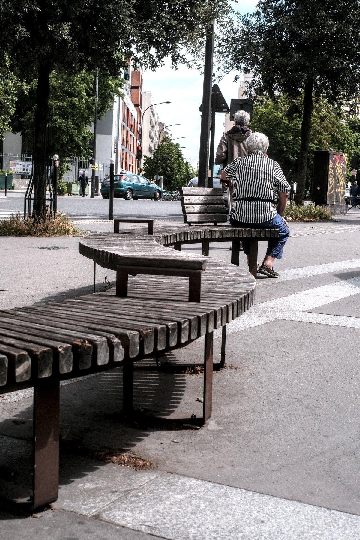 Photo verticale. Sur une place publique, un banc fait de petite lattes de bois assemblées sur leur tranche et de manière à serpenter. Une partie posée dessus apparaît à un moment serpentant dans l’autre sens et prolonge le banc tout en restant bien à plat, la place étant penchée. À l’extrémité, une dame au cheveux blanc et courts, avec un haut à rayure verticales noires et blanches est assise