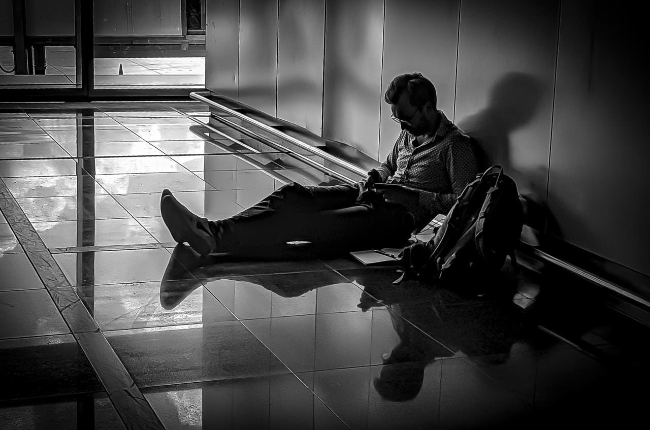 Man sitting on a polished floor, leaning against a wall, engrossed in a device. Backpack and papers rest beside him, while his shadow is cast on the wall. Sunlight streams through a glass door, creating reflections on the tiled floor. Image in black and white, emphasizing contrast and shadows.
