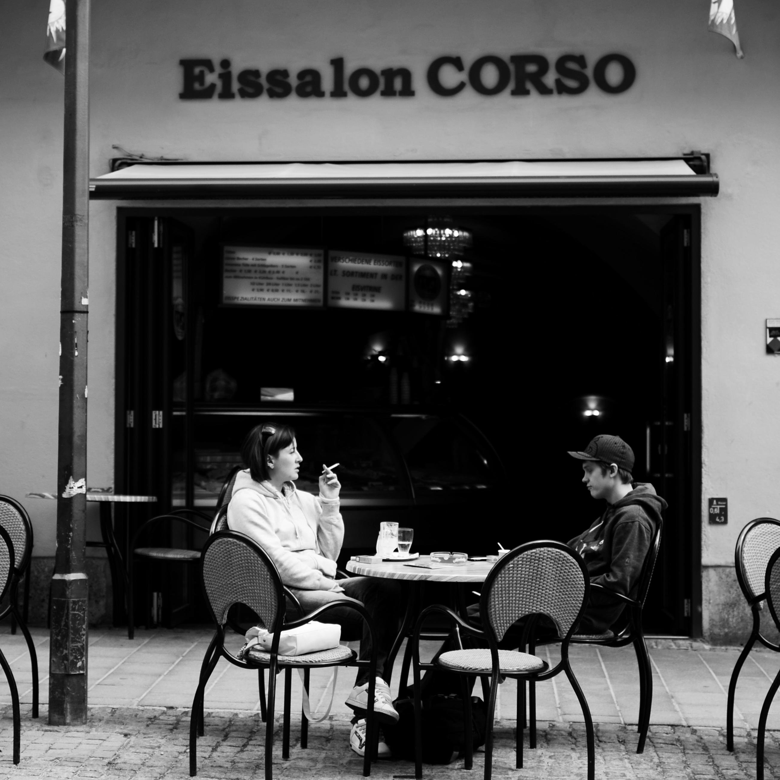 Schwarz-Weiß-Straßenfoto von zwei Personen, die an einem Café-Tisch im Freien vor einem Schaufenster mit der Aufschrift „Eissalon CORSO“ sitzen, mit leeren Stühlen um sie herum und einem Getränk und kleinen Gegenständen auf dem Tisch.