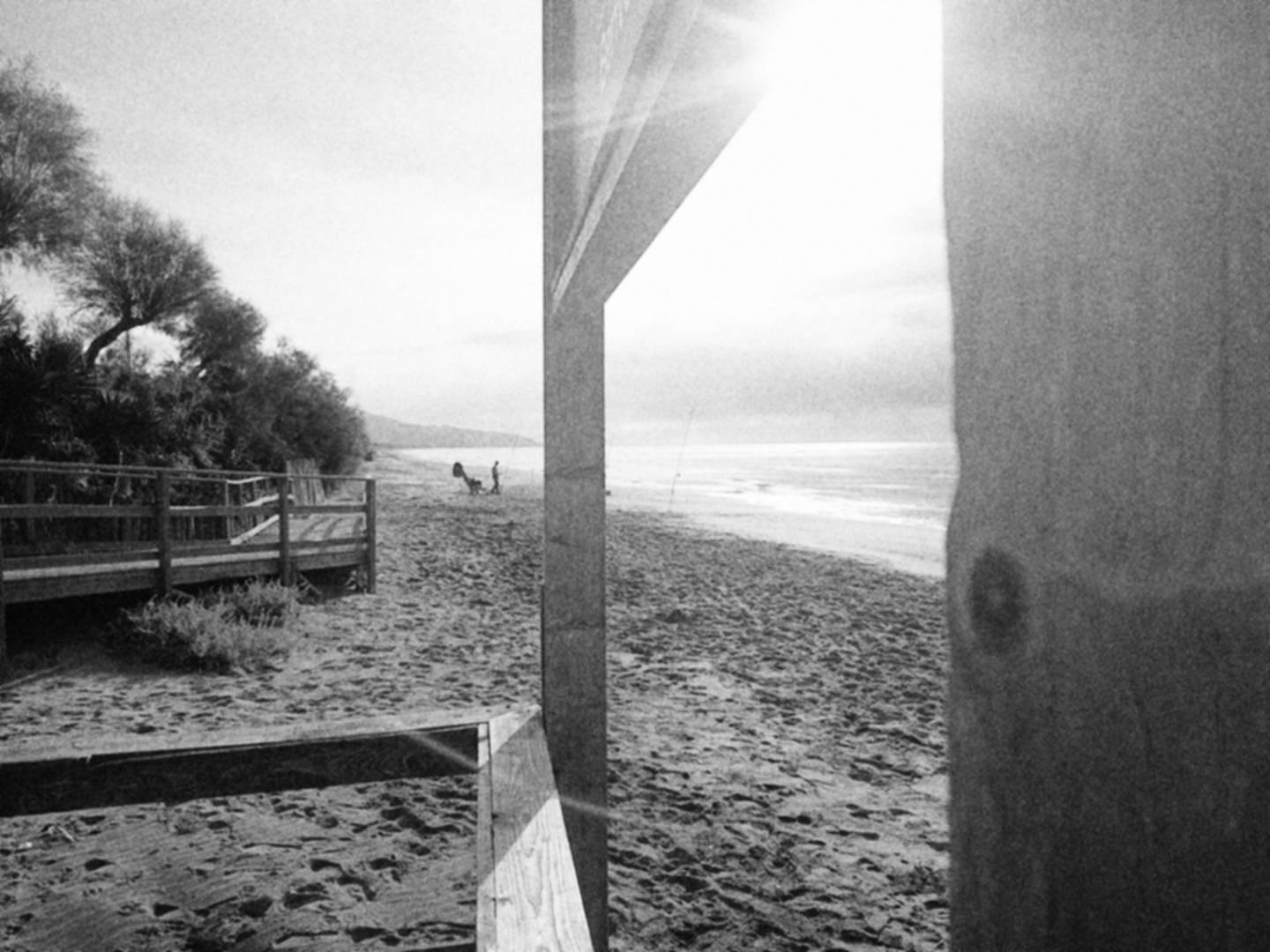 Spiaggia con arbusti tipici sulla sinistra e un pescatore, quasi in silhouette, con la sua sedia a sdraio e le canne da pesca piantate nella sabbia. Il cielo è nuvoloso e la foto in bianco e nero è scattata attraverso una struttura in legno.