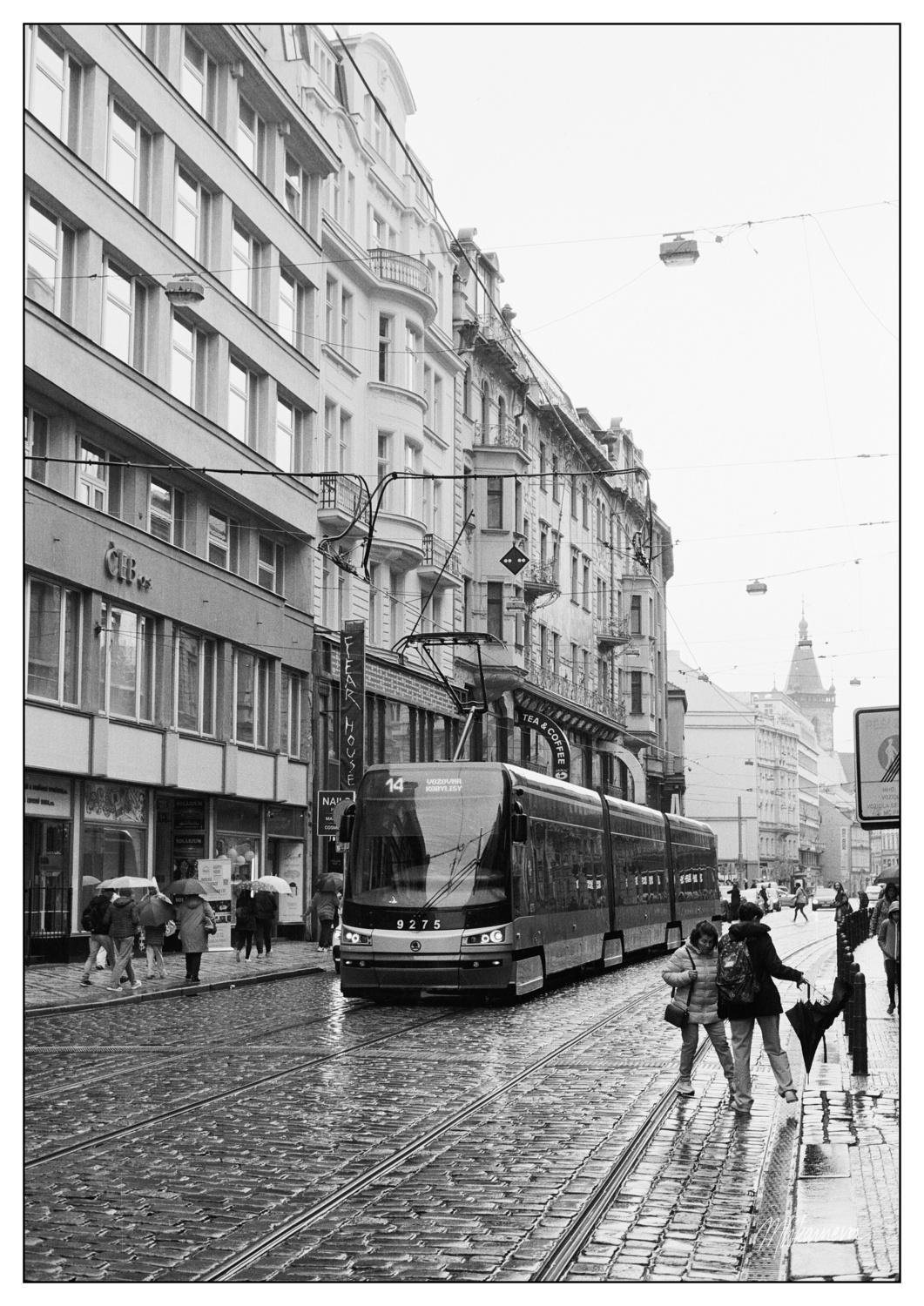 Black and white image of a city street with a modern tram numbered 9275. The tram is on a cobblestone street lined with tall, historic buildings. Pedestrians with umbrellas walk along the wet sidewalks, indicating recent rain. Overhead wires are visible, connecting to the tram. In the background, there is a distant view of a tower.