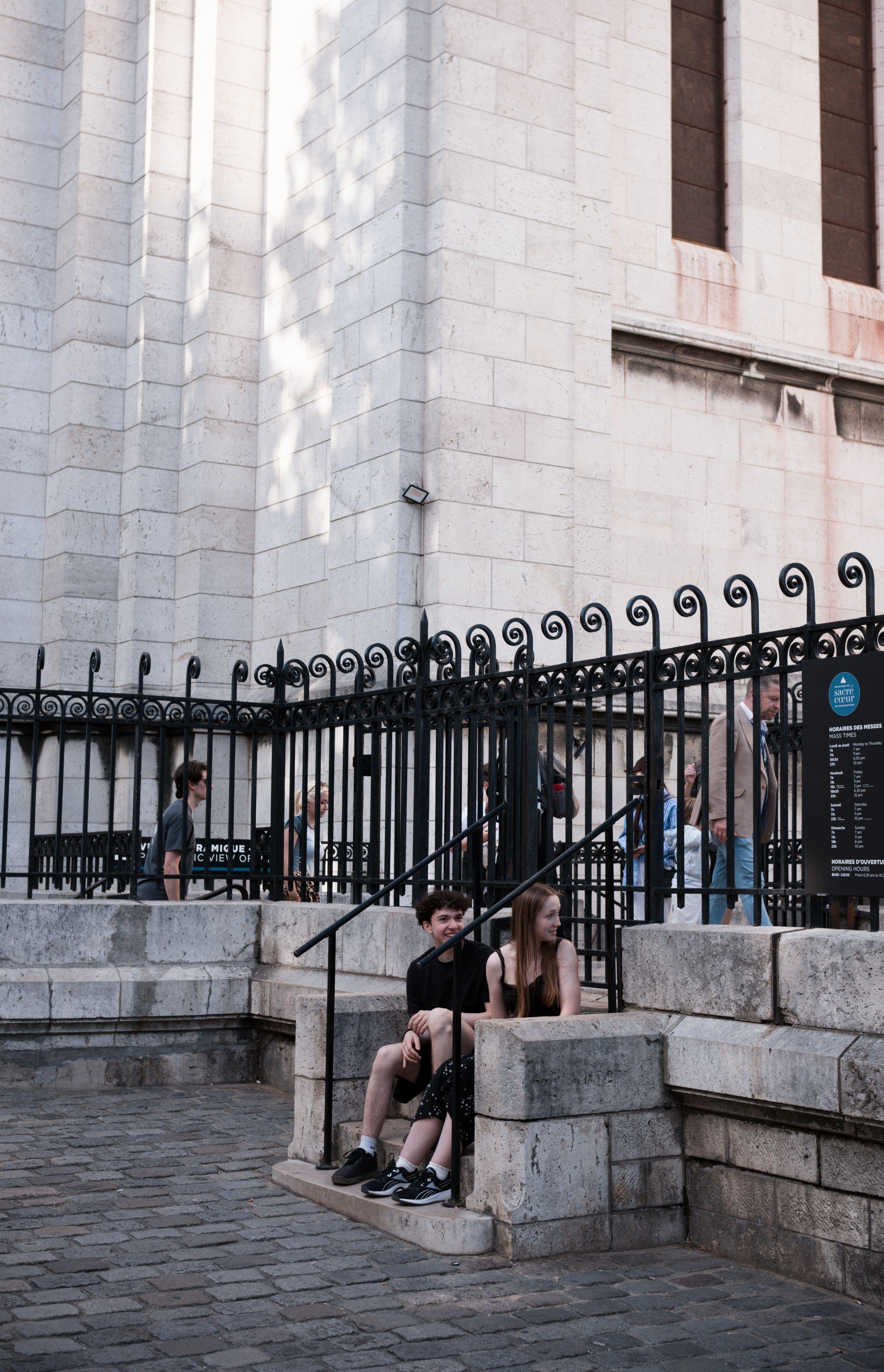 Photo au format portrait de 2 adolescents assis sur un petit escalier en pierre. Une clôture noire est derrière eux au-dessus de l'escalier d’environ 5 marches, murs blancs du Sacré Cœur derrière la clôture. La fille aux longs cheveux teints dans un roux foncé tourne sa tête derrière elle pour regarder quelqu'un, le garçon aux cheveux noirs courts tout bouclés, regardait également mais s’est aperçu de ma présence et tourne son visage vers la caméra. Derrière la clôture, certains touristes marchent vers la droite de la photo