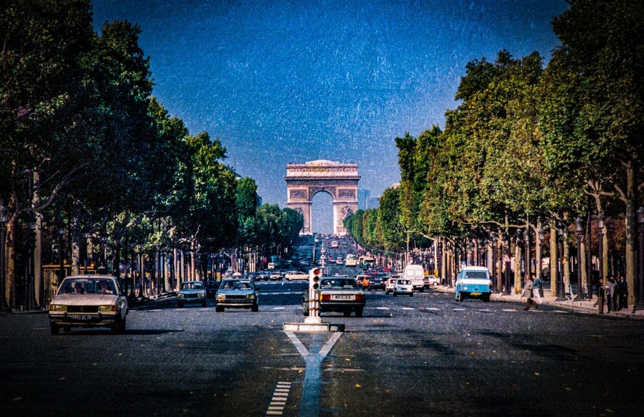 Cars driving along a wide, tree-lined avenue with the Arc de Triomphe in the center background. The road is busy with vehicles, and the sky is clear and blue. The scene has a vintage appearance with a textured look.