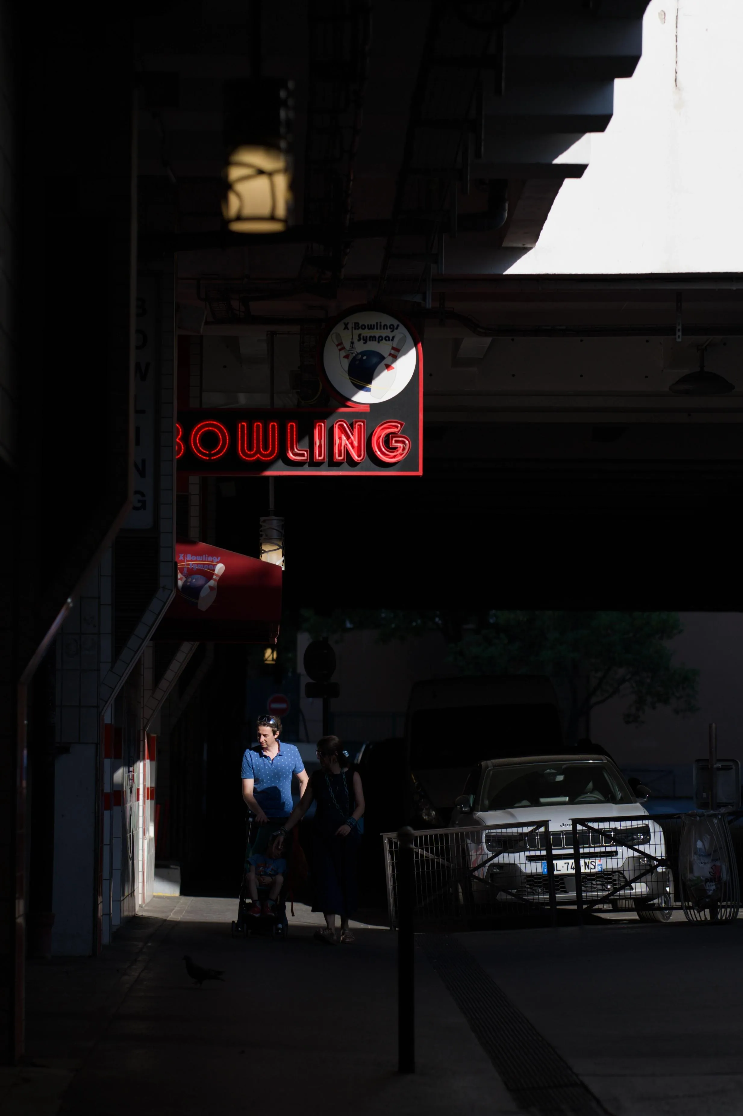 Photograph of three people walking on a street sidewalk towards the camera, under a sign for a bowling place. The three people visible are a man, a woman by his side, and a child in a stroller the man is pushing. The covered street is nearly all dark, except for a light ray coming diagonally from an opening in the roof. The sun ray is hitting part of the bowling sign as well as the man's torso and head, but the stroller and the woman are in the dark.
