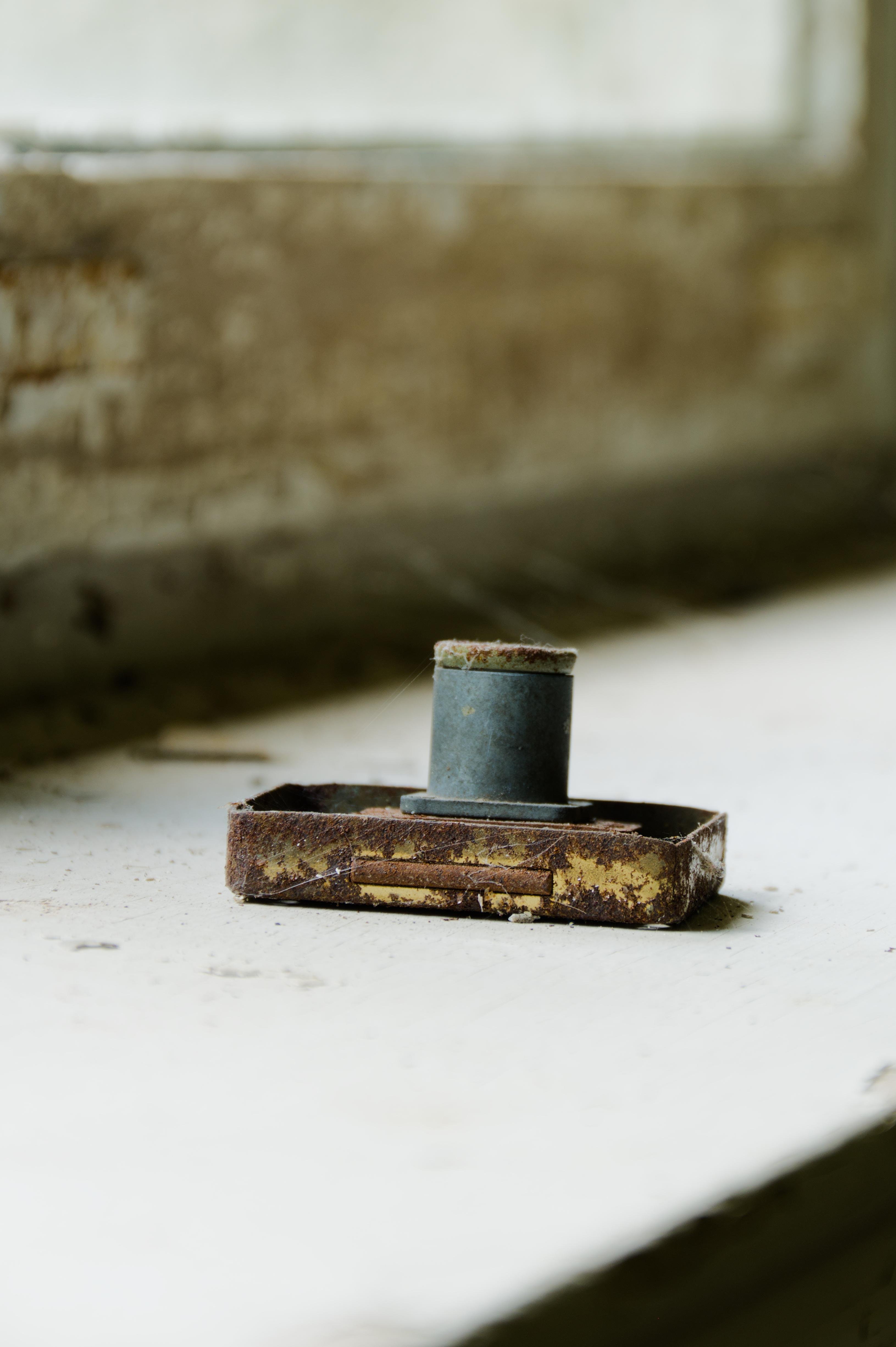 A rusty, old furniture lock lies on a dusty windowsill. In the background, a blurry view of a stained, peeling window frame can be seen. 
###
Eine rostiges, altes Möbelschloss liegt  auf einem staubigen Fensterbrett. Im Hintergrund ist ein verschwommener Blick auf einen fleckigen, abblätternden Fensterrahmen zu sehen.