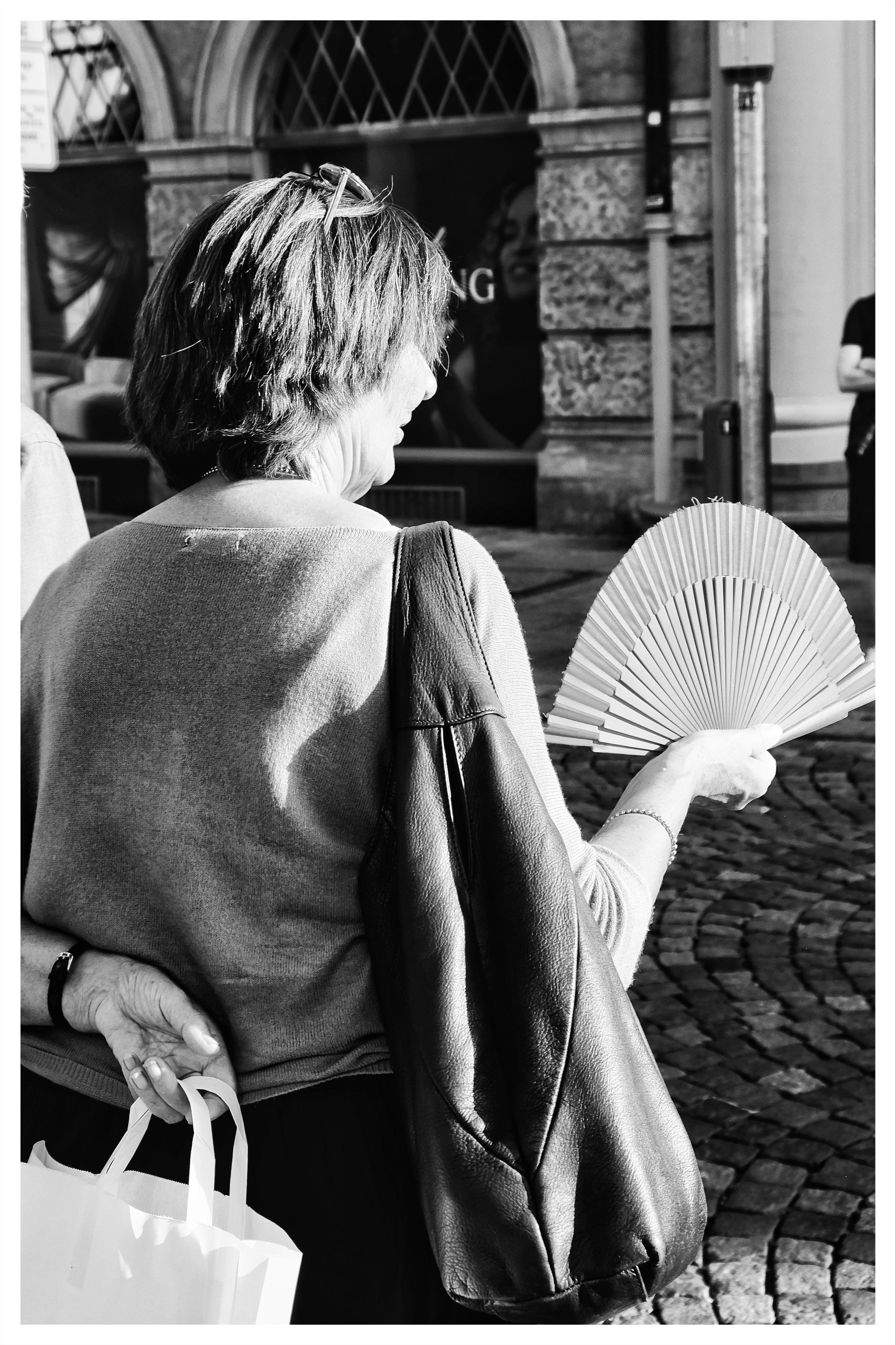 Black-and-white street photo of a woman seen from behind holding an open folding fan, with a large shoulder bag on her arm and shopping bags in her other hand, standing on a cobblestone street near a stone building with arched windows.
