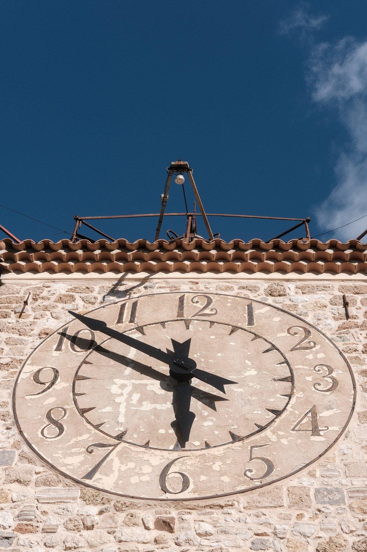 Photo verticale. Gros plan sur une horloge dans un mur de pierre. Au dessus des bouts de tuile apparaissent, puis une structure métallique pour porter de l’éclairage et enfin, le ciel bleu profond, avec un bout de nuage à droite