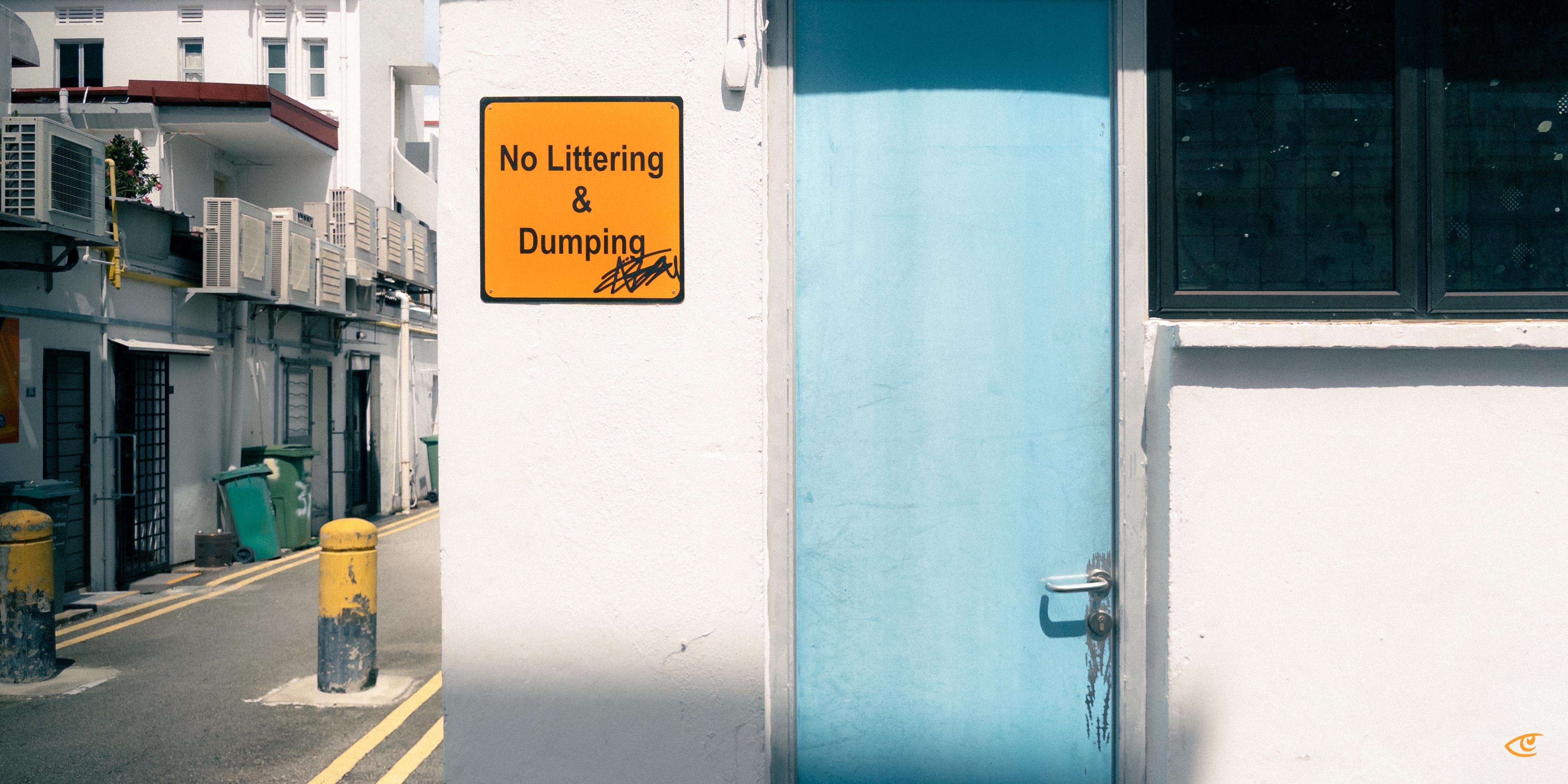 Orange “No Littering & Dumping” sign on a white wall beside a light blue door with a metal handle; narrow alley to the left with yellow bollards, trash bins, and building air-conditioning units.