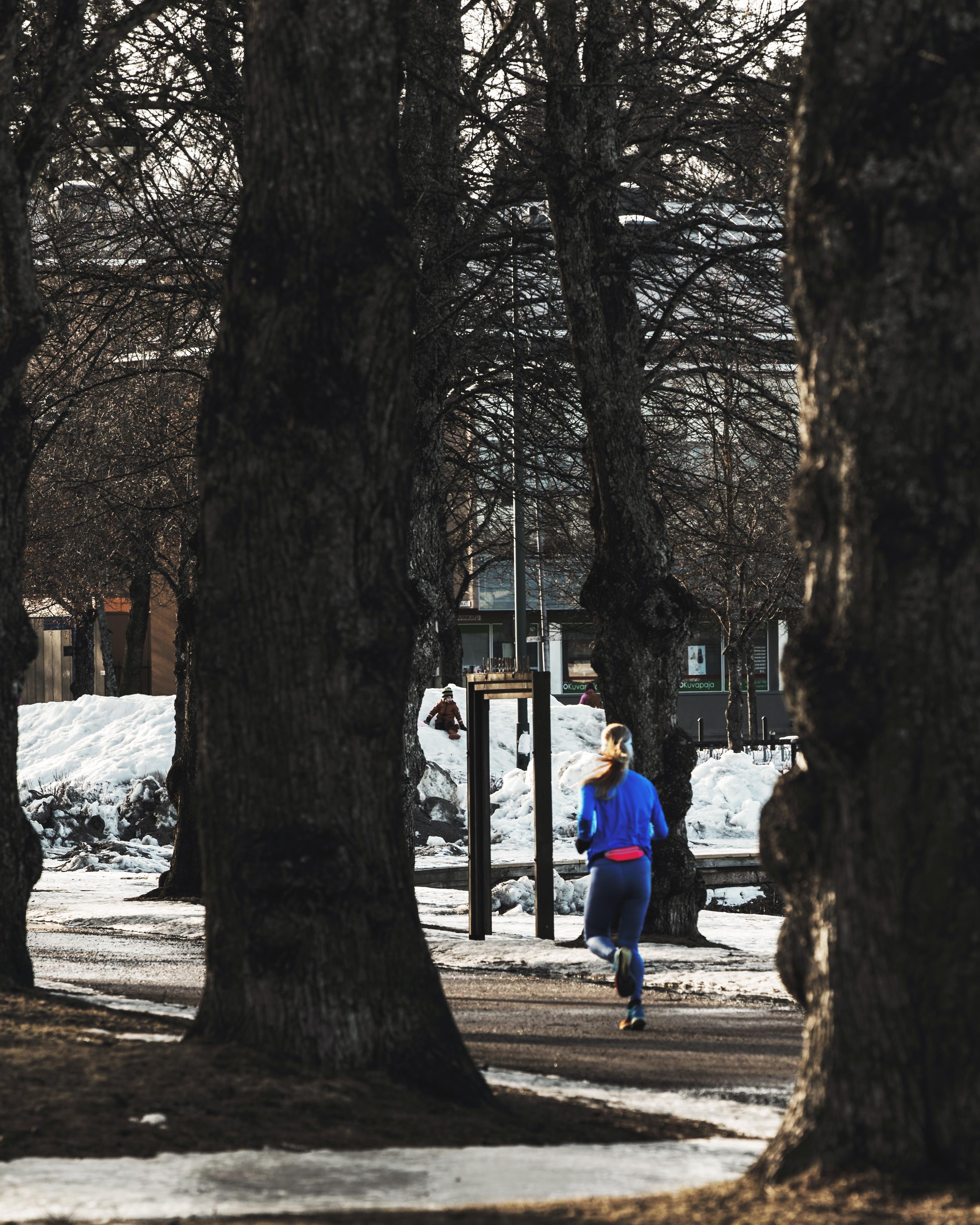 The image shows a person jogging in a park area. The person is wearing a blue jacket and there's snow on the ground. The scene is framed by bare trees, suggesting it's a colder season. A signpost and benches are visible in the background.