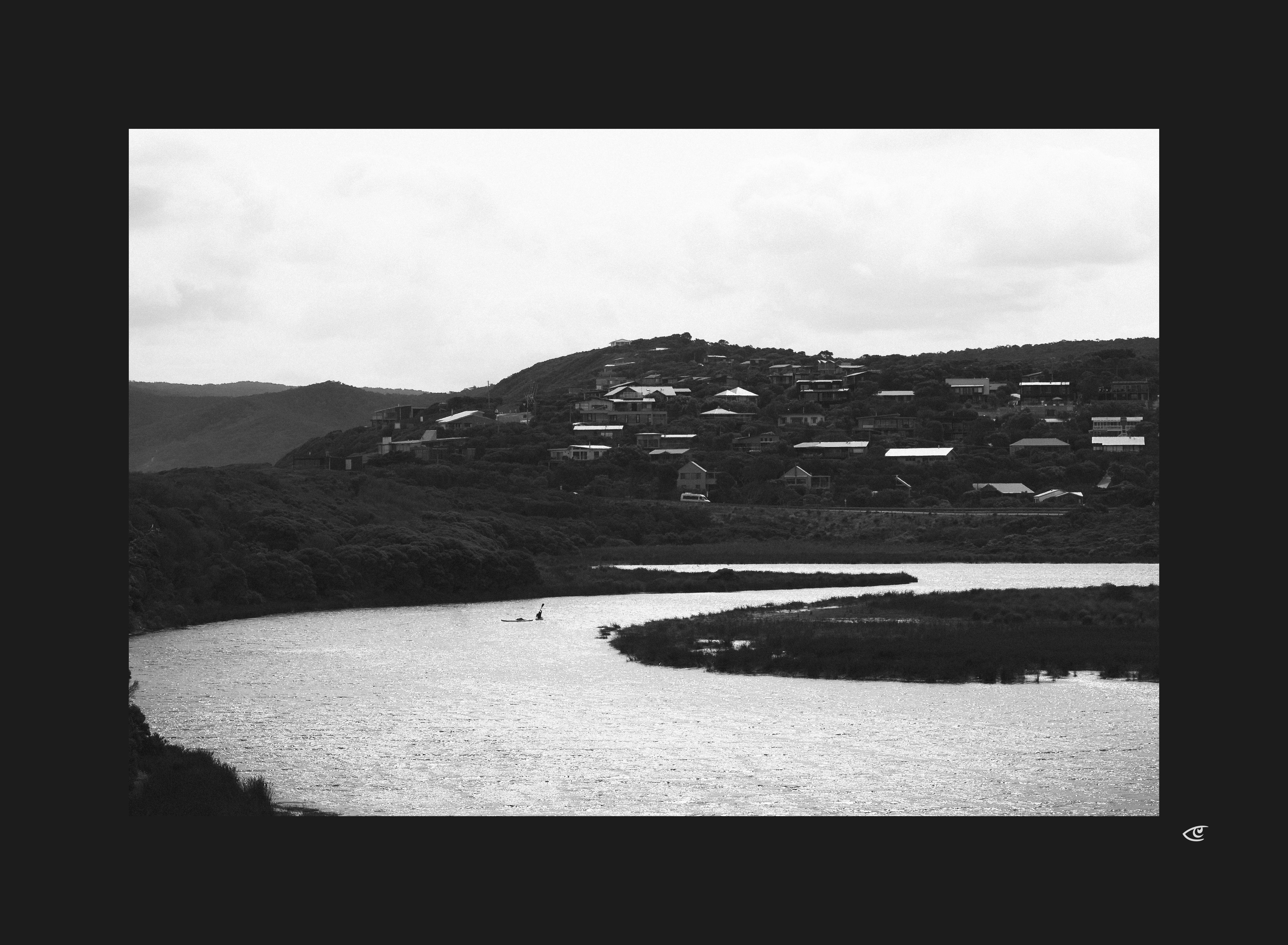 Black-and-white landscape of a winding river in the foreground, with marshy banks and a small canoe on the water, leading to a hillside settlement of scattered houses under a cloudy sky.