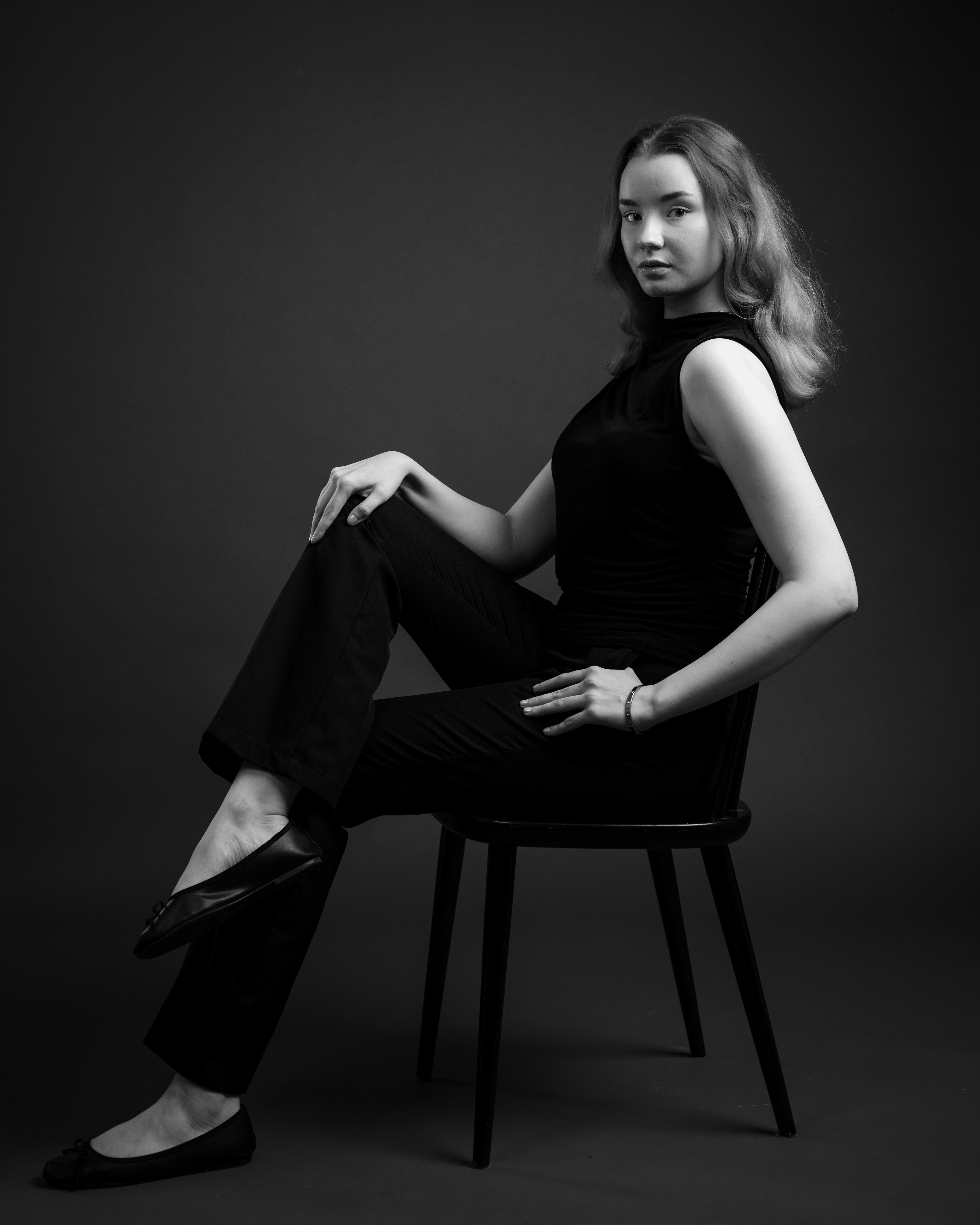 This is a black and white portrait of a person sitting on a chair. The person is wearing dark clothing and has long hair. Their pose is relaxed, with one leg crossed over the other. The background is plain, adding emphasis to the subject. Mallikuvaus. Mallikansio muotikuvaaja Kuvaajankulma