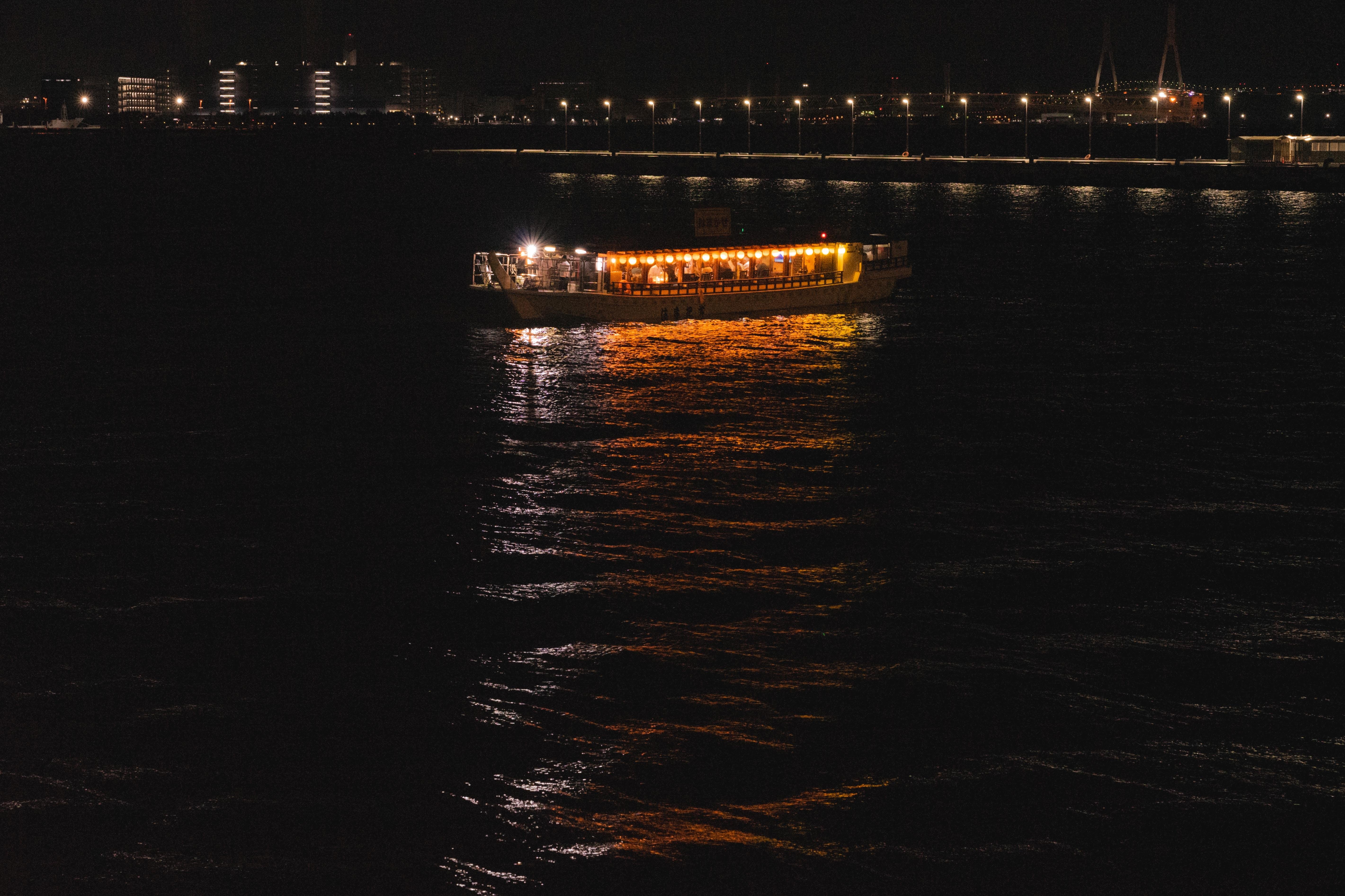 A boat brightly lit with orange and yellow lights travels across a dark body of water at night. The lights reflect on the water's surface, creating a glowing path. In the background, urban buildings and streetlights are visible along the shoreline.