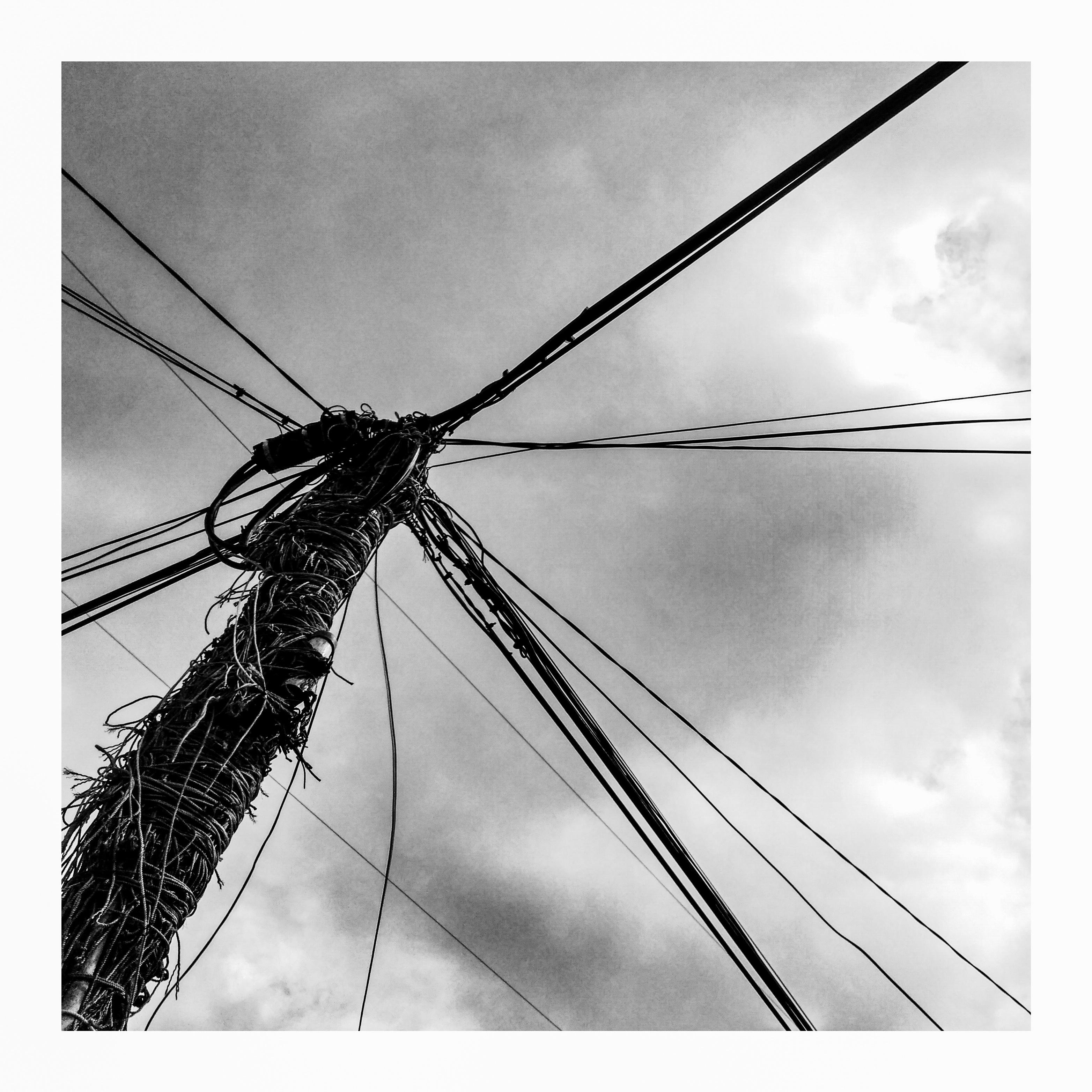 Low-angle black-and-white view of a utility pole with tangled wiring and multiple cables radiating outward against a cloudy sky.