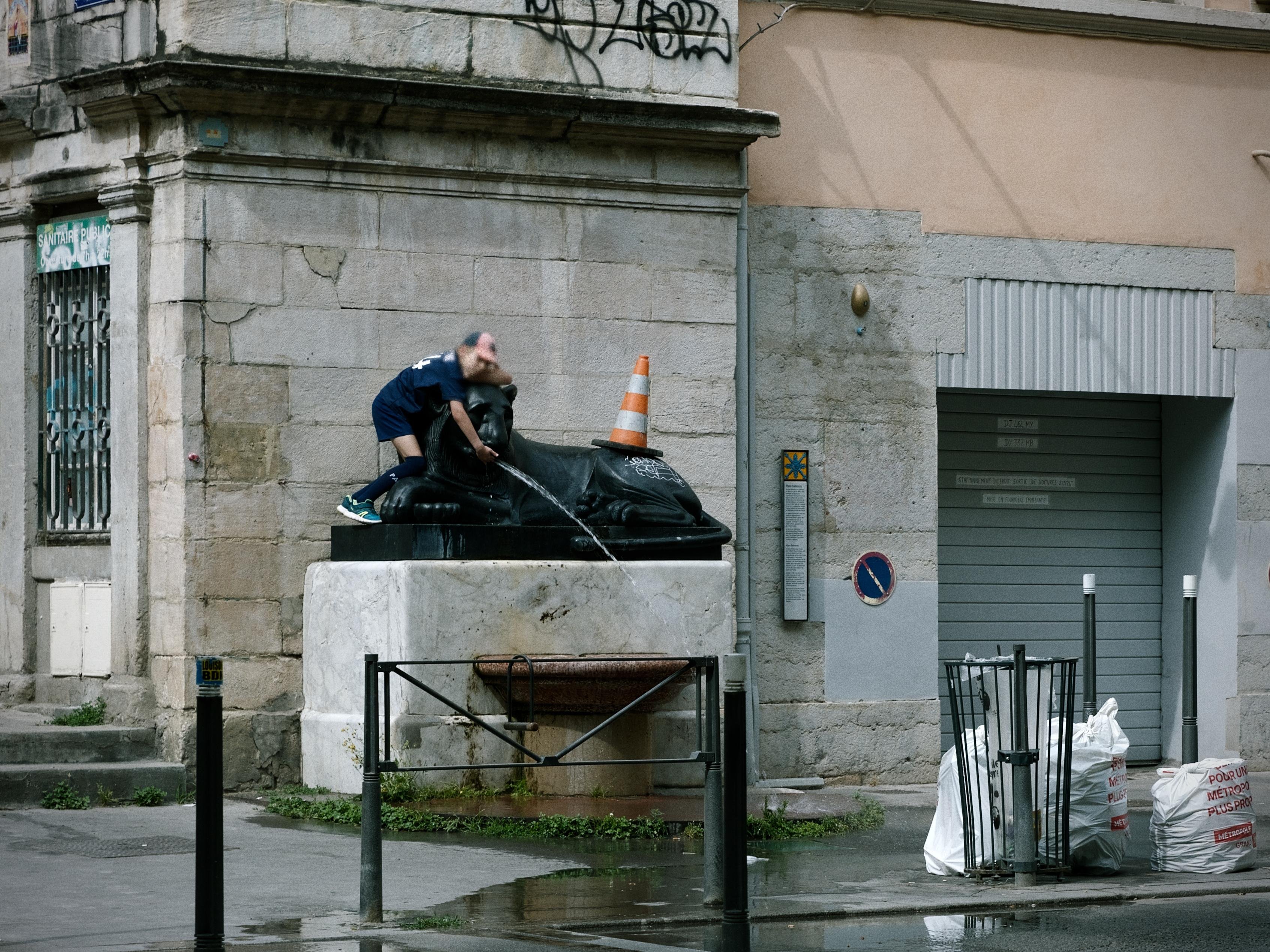 A young boy standing upon a sculpted fountain that looks like some kind of feline. The surrounding area is a messy-looking street with a garage door, wet and dirty concrete bricks, bags of cement and a traffic cone lying atop the fountain next to the boy. He is using his hand to alter the direction of the fountain stream towards the pavement in a very evocative manner. The overall scene has a nostalgic, film-like atmosphere, emphasised by the grain, slight vignetting and the greenish white balance. The action of the boy is central to the storytelling, playfully ignoring rules.