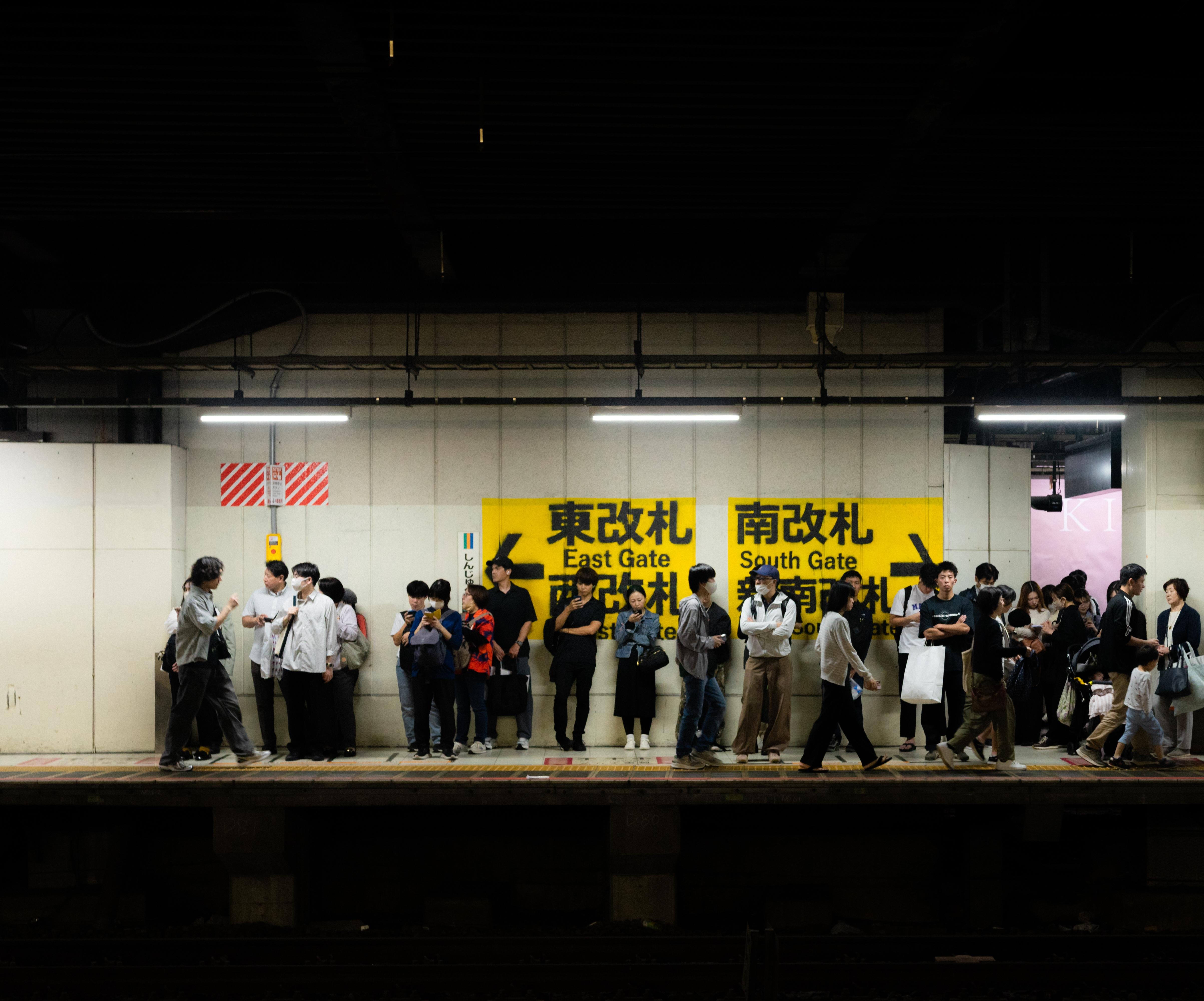 People standing on a subway platform waiting for a train, viewed under bright overhead lights. Yellow signs with black text indicate directions to East Gate and South Gate. The setting appears to be an urban transit station, with individuals of varying ages and attire, some interacting or using phones.