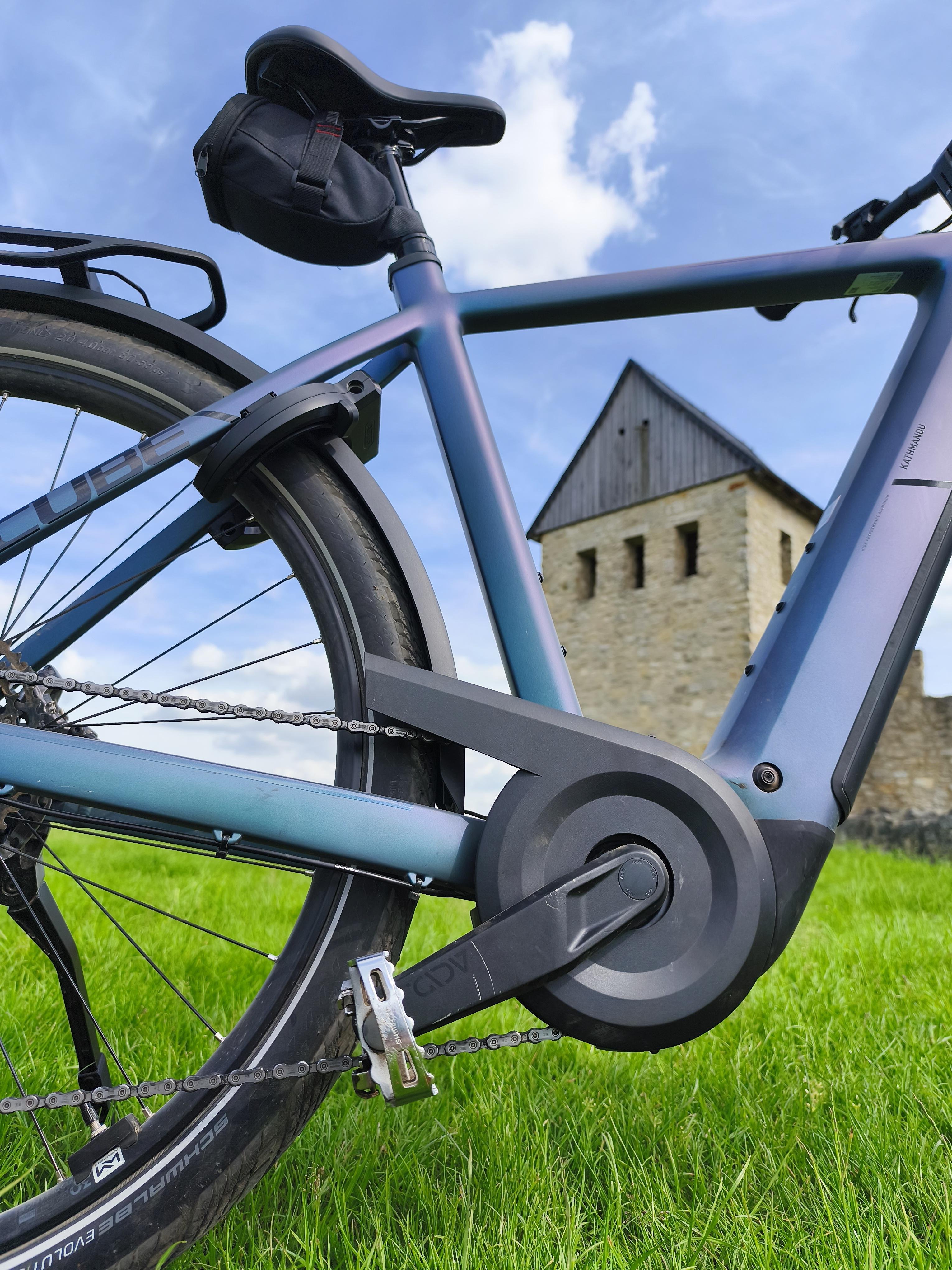 Close-up, low-angle view of an electric bicycle’s rear wheel, chain, and mid-drive motor with saddle and small under-seat bag, set on green grass with a stone tower in the background under a blue sky with clouds.