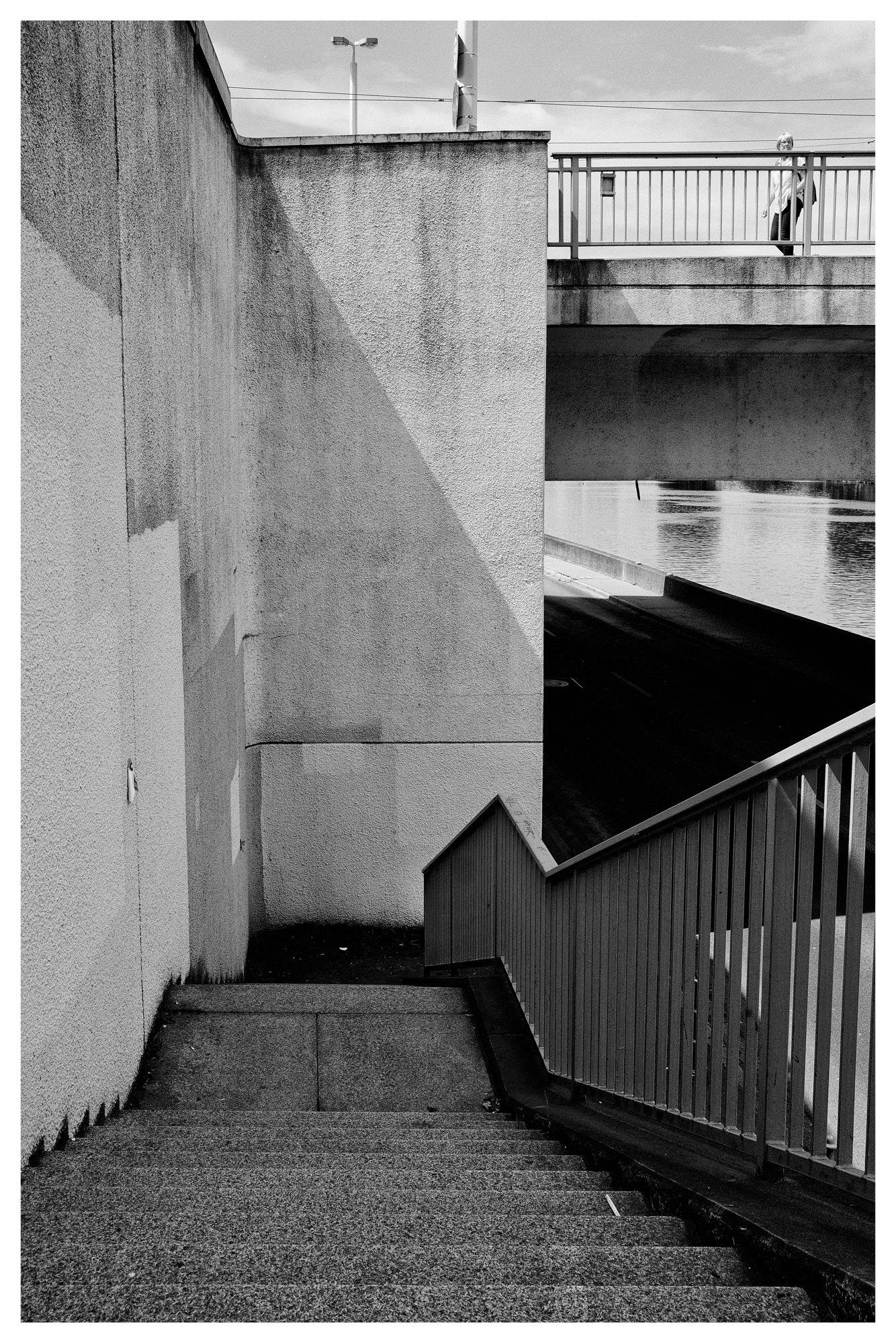 Black-and-white photo of a concrete stairway descending beside a tall wall, with a metal railing on the right leading toward a canal underpass; a pedestrian walks across a bridge at the top right above the water.