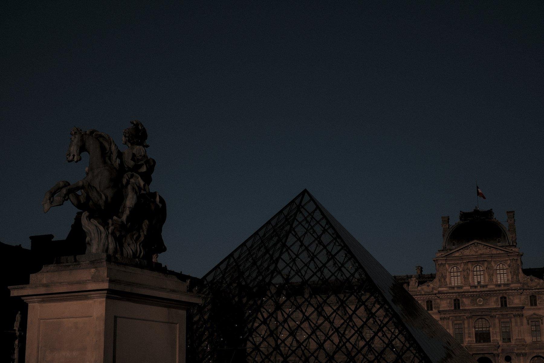 Photo horizontale. On voit la statue de Louis XIV, puis la Pyramide du Louvre et le Louvre sur 3 plan différent, en diagonale, dans une ambiance feutrée dû à l’éclipse