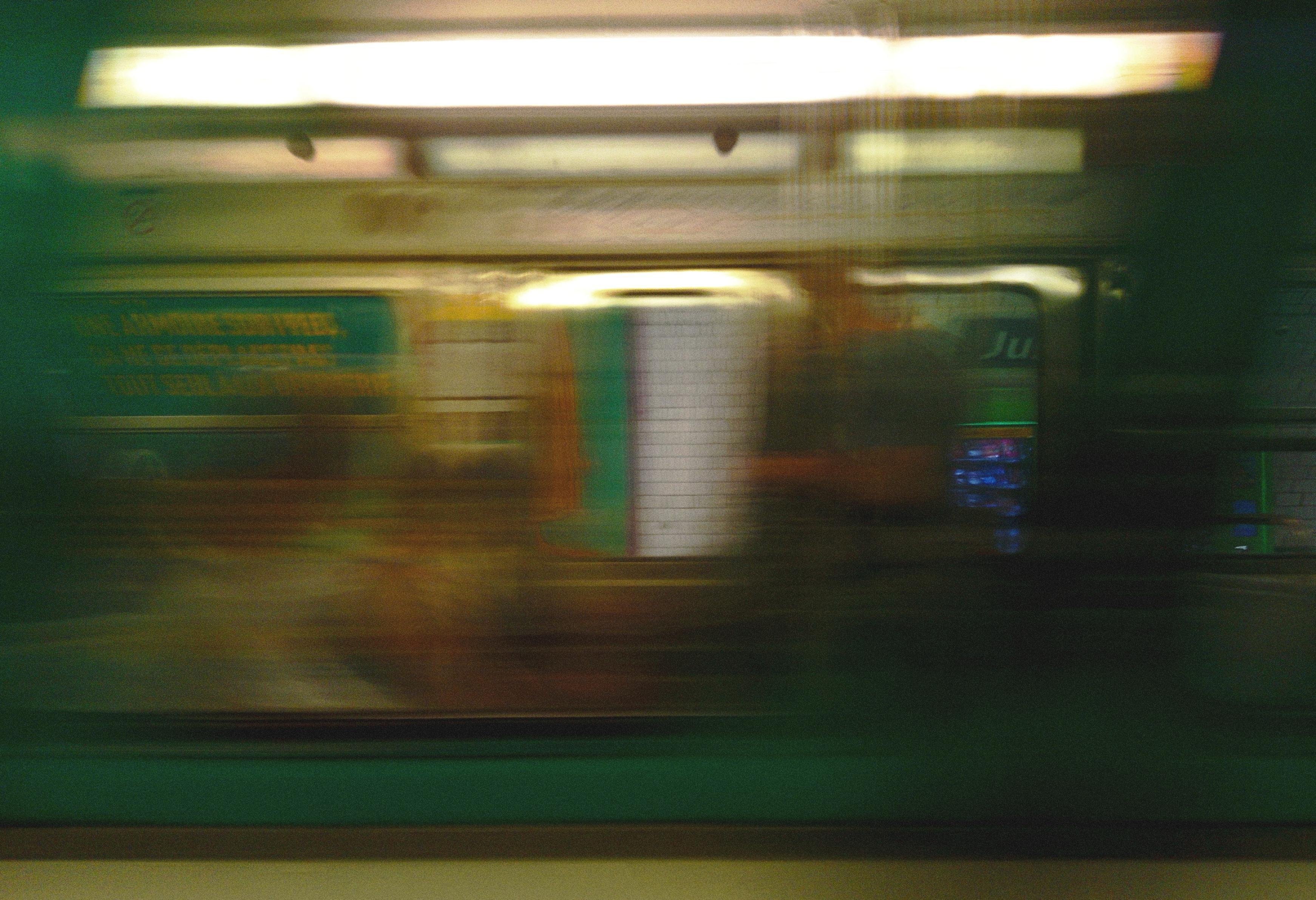 Foto hecha desde un metro parisino en movimiento, viéndose el interior de otro metro en movimiento y la zona peatonal que hay detrás, también en "movimiento". La iluminación es tenue y verdosa/amarillenta por el color del metro