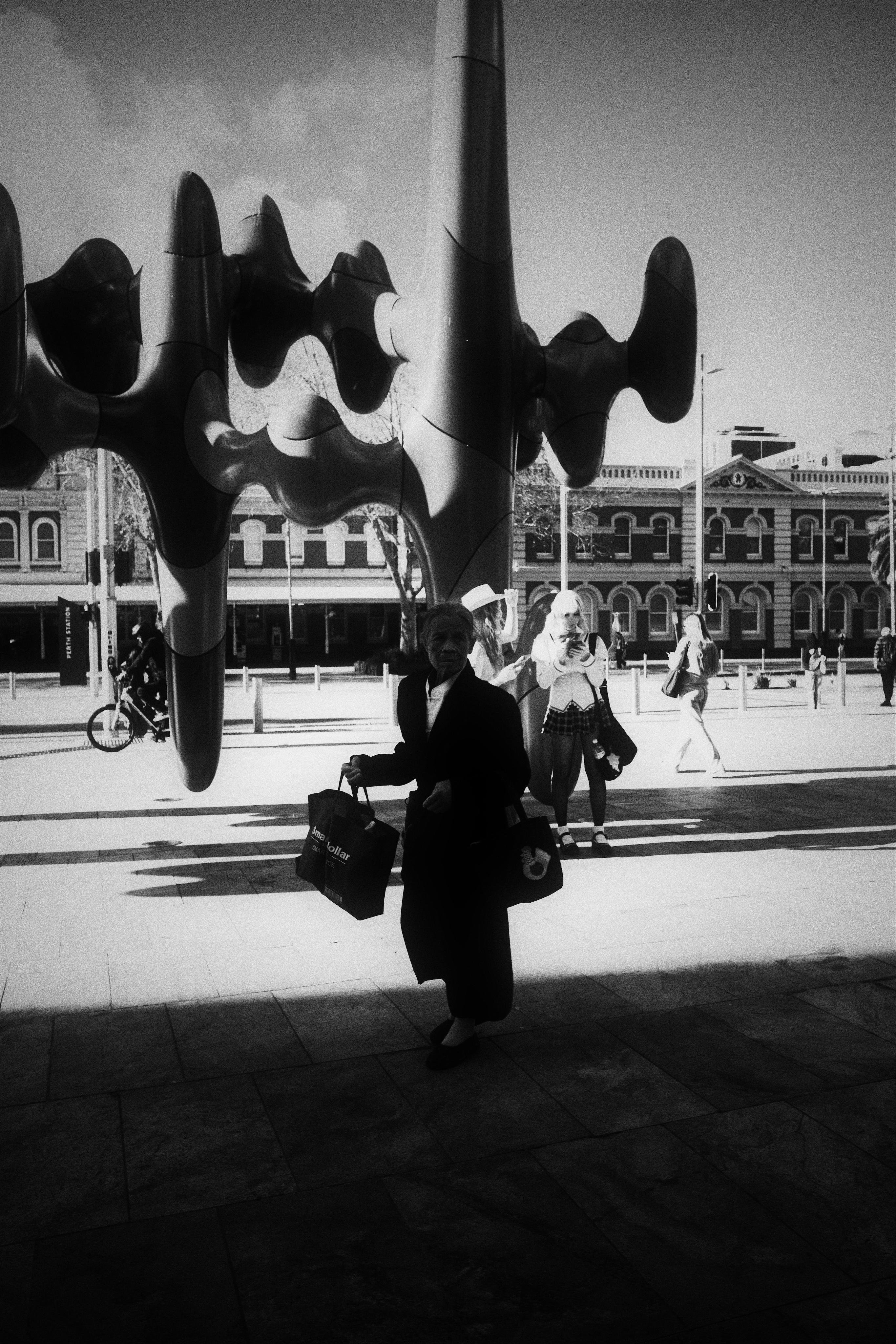 Black-and-white street scene with a large abstract metal sculpture of curved, bulbous forms overhead; a silhouetted person carrying shopping bags walks in the foreground while pedestrians and a cyclist pass in front of low historic buildings across the street.