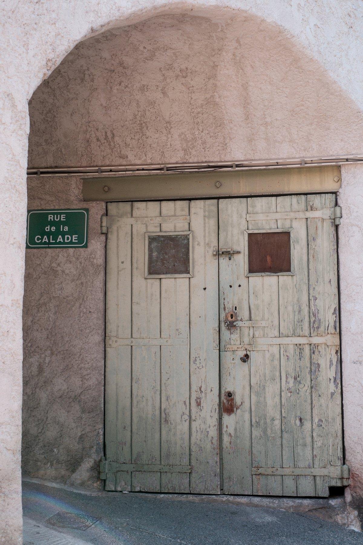 Photo verticale. Sous l’entrée d’un tunnel piéton rose, une double porte en bois peinte d’une couleur probablement claire, mais patinée par le temps, donnant du beige au gris. Sur chaque volant de porte, une petite fenêtre carrée en haut