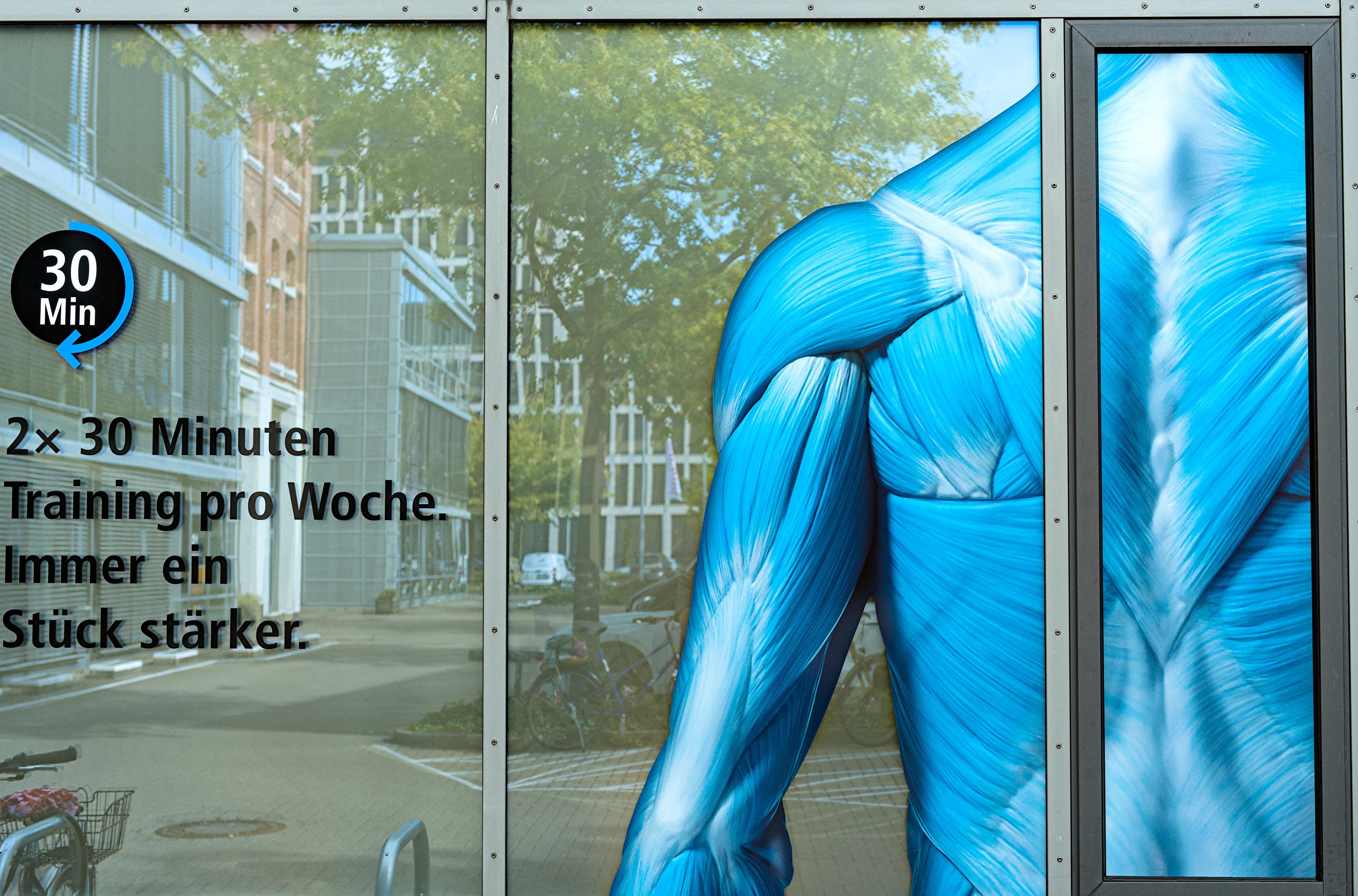 Blue anatomical model of a muscular upper back and arm displayed behind a glass window. German text on the left reads "2× 30 Minuten Training pro Woche. Immer ein Stück stärker," with an icon showing "30 Min" highlighted in a black circle. Building and trees are faintly reflected in the glass, indicating an urban setting.
###
Blaues anatomisches Modell eines muskulösen oberen Rückens und Arms, das hinter einem Glasfenster angezeigt wird. Der deutsche Text lautet "2 x 30 Minuten Training pro Woche. Immer ein Stück stärker", mit einer Ikone, die "30 Min" zeigt, die im schwarzen Kreis hervorgehoben wird. Gebäude und Bäume spiegeln sich schwach im Glas wider, was auf eine urbane Umgebung hinweist.