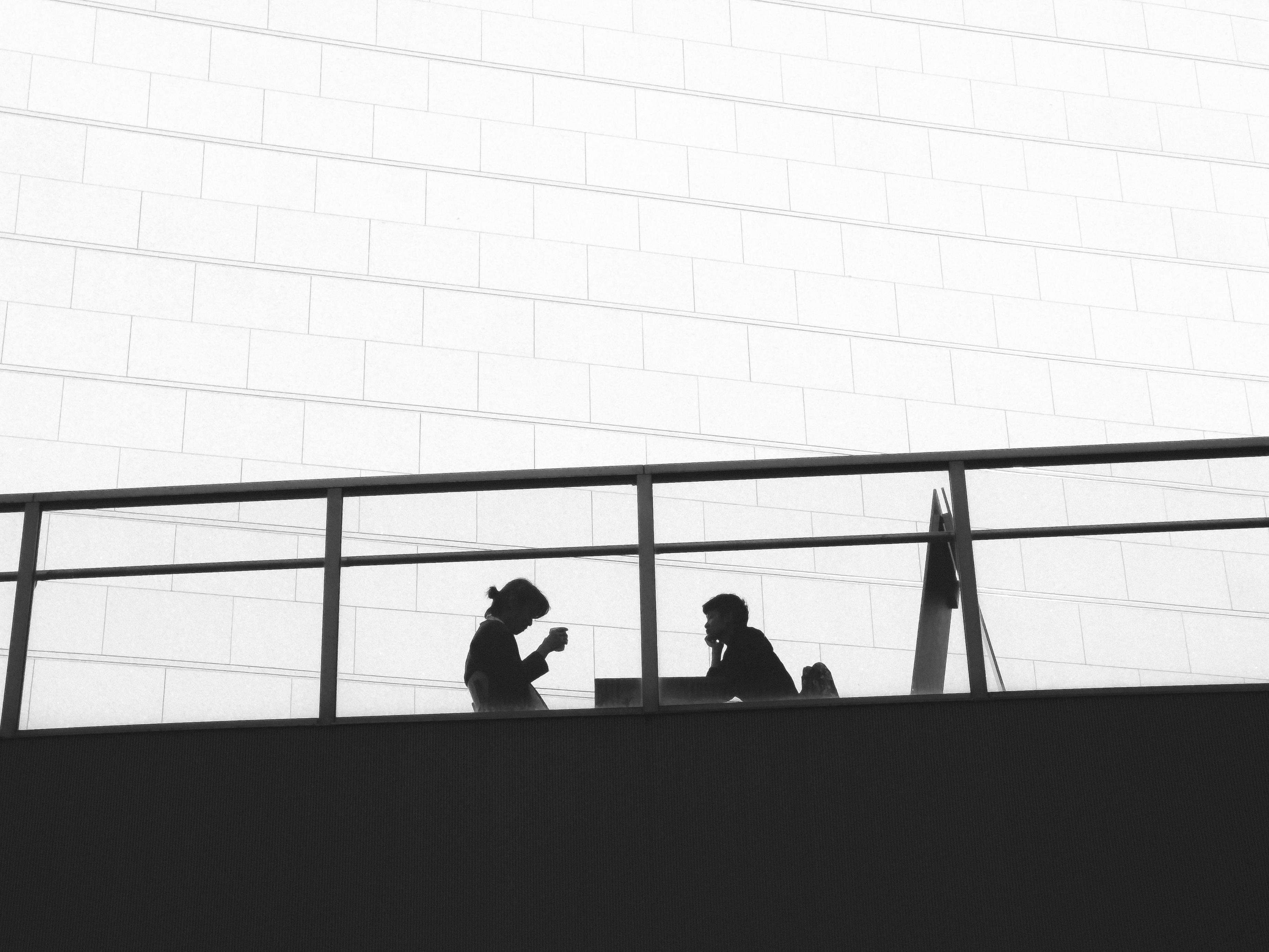 This black and white image shows the silhouettes of two people sitting at a table, possibly having a conversation. One person appears to be holding a cup. They are on a balcony or terrace with a geometric, tiled wall as a backdrop. The image conveys a sense of minimalism and modern architecture.