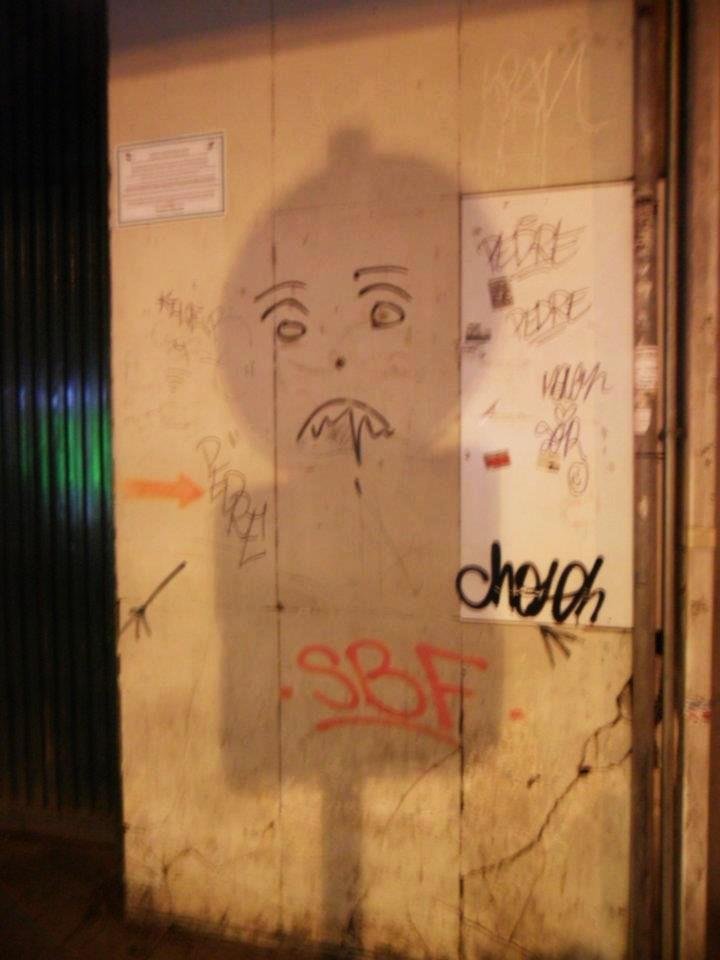 Fotografía de una pared en una calle de una ciudad. Es de noche y se proyecta la sombra de un poste que tiene una señal de tráfico cuadrada y encima otra redonda. En la pared hay pintada una cara con ojos, cejas y una boca seria que de la que parece que cae saliva o algún otro líquido. Hay otros graffitis en la pared.
