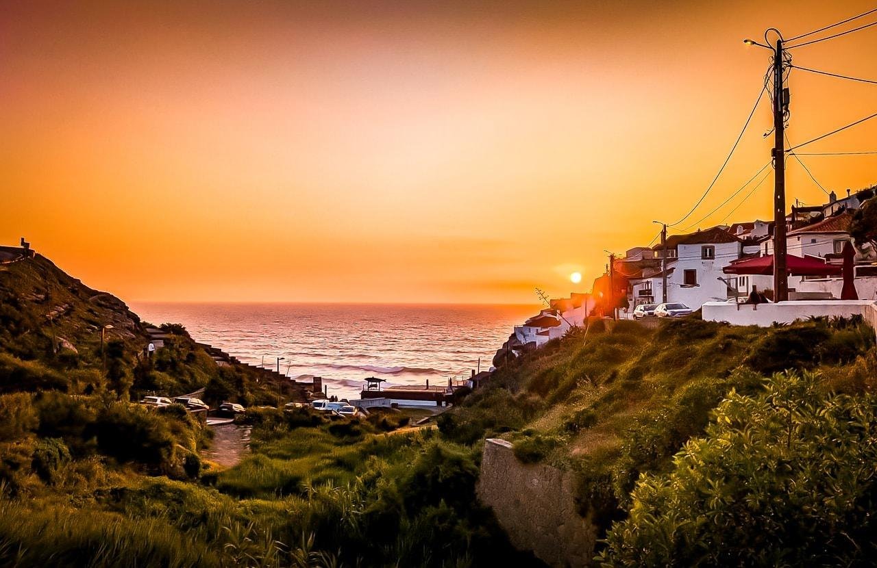 Sunset over a coastal village with orange and red hues in the sky. The sun is setting on the horizon above a calm ocean. White houses with red roofs line the right side, perched on a grassy hill. Power lines and a few parked cars are visible alongside narrow roads.