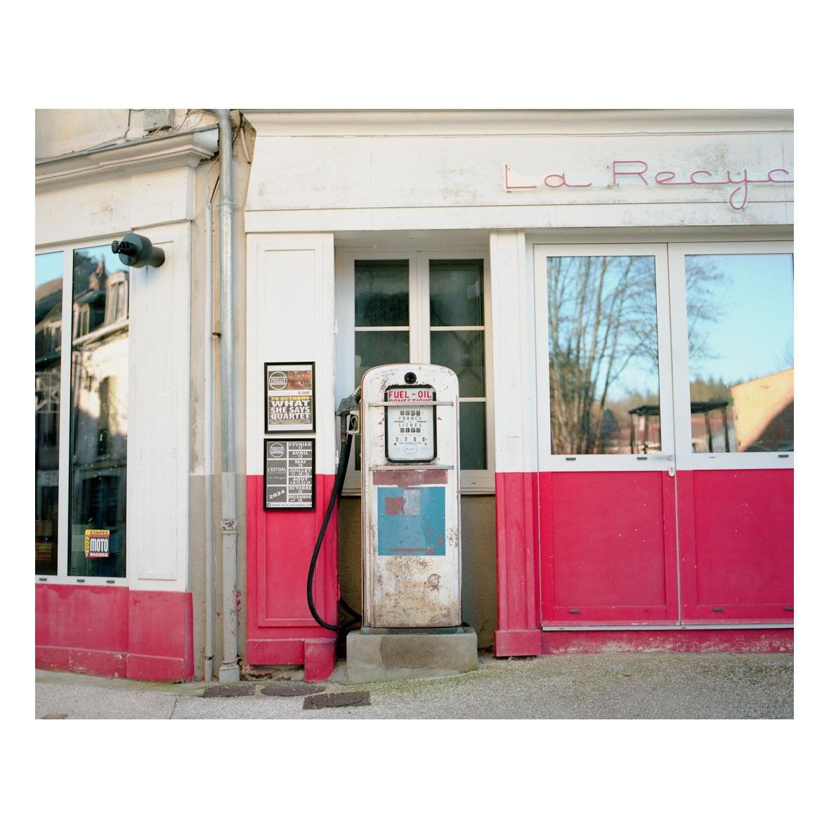 The image depicts an old-fashioned fuel pump in front of a building, possibly repurposed or abandoned. The building has a red and white color scheme, and the word "La Recyclerie" is partially visible.